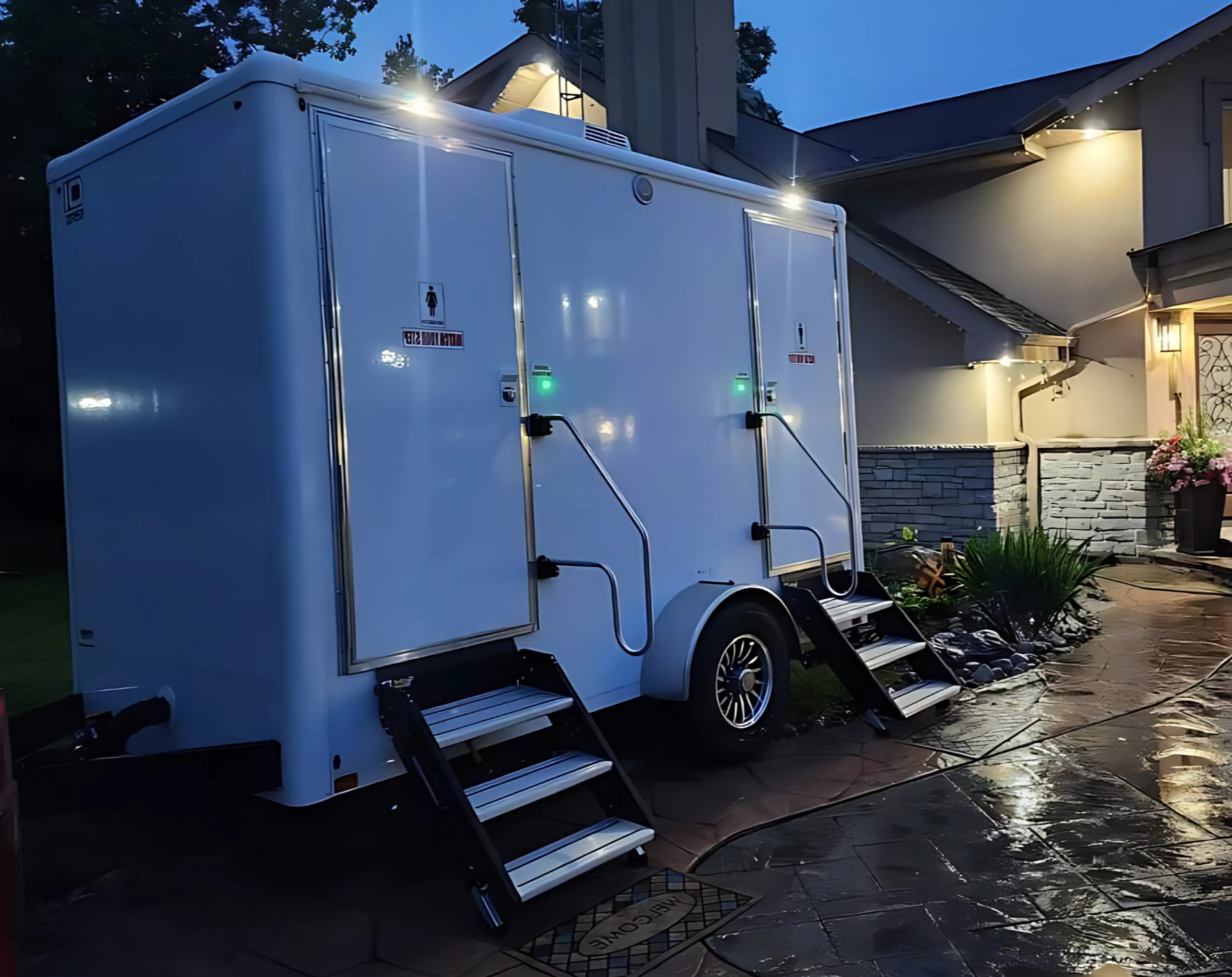 White mobile restroom trailer with stairs and handrails, set up outside a house in evening light.
