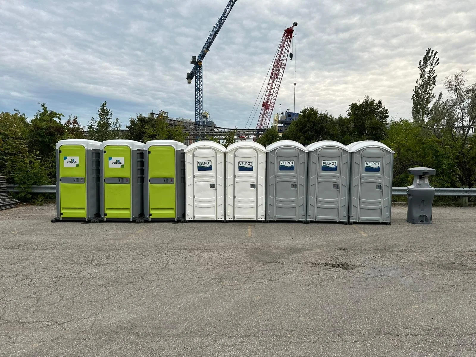 A row of green, white, and gray portable toilets on a paved surface with construction cranes and greenery in the background.