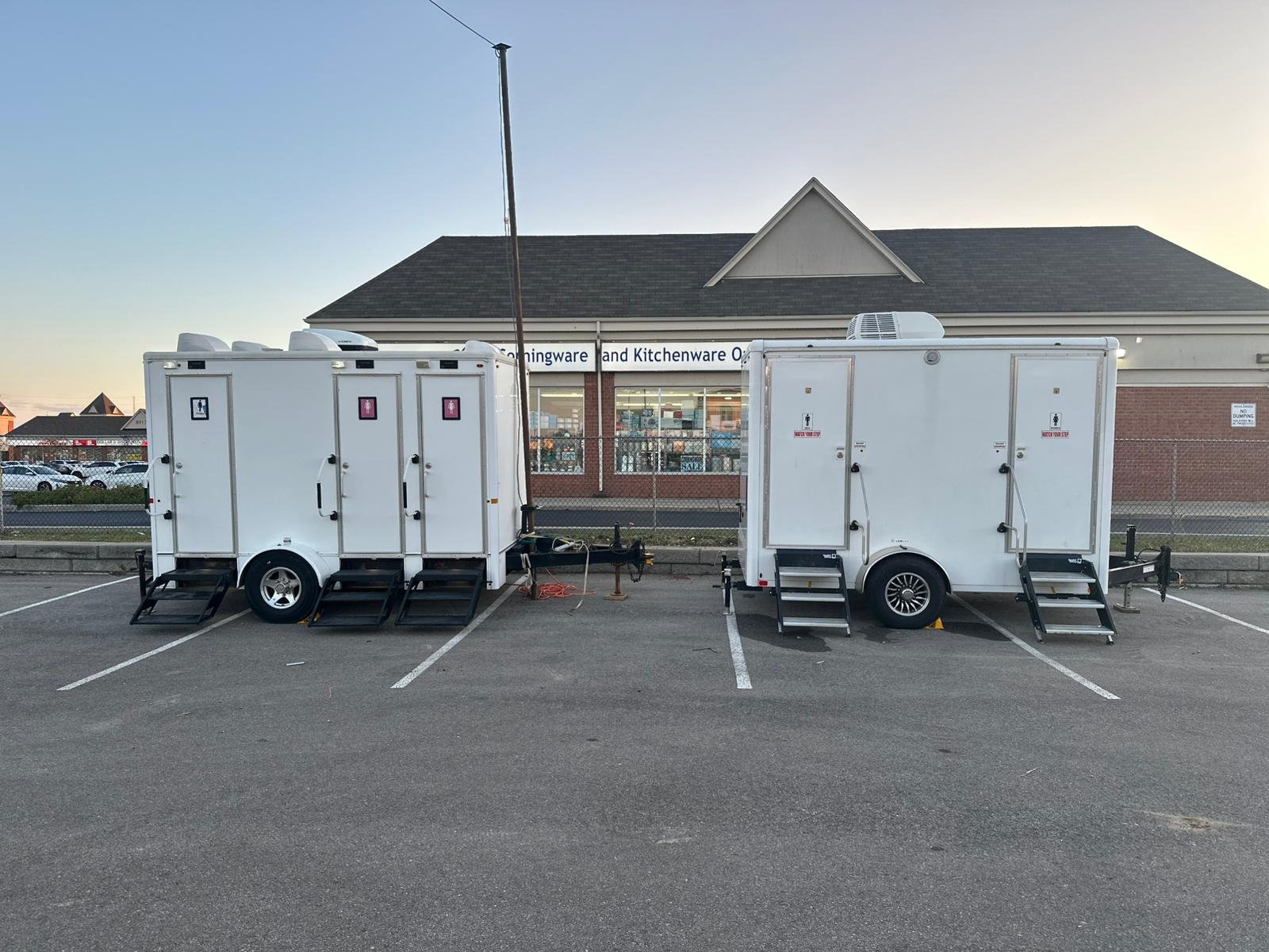 Two white portable restroom trailers parked in a commercial parking lot with a store in the background.