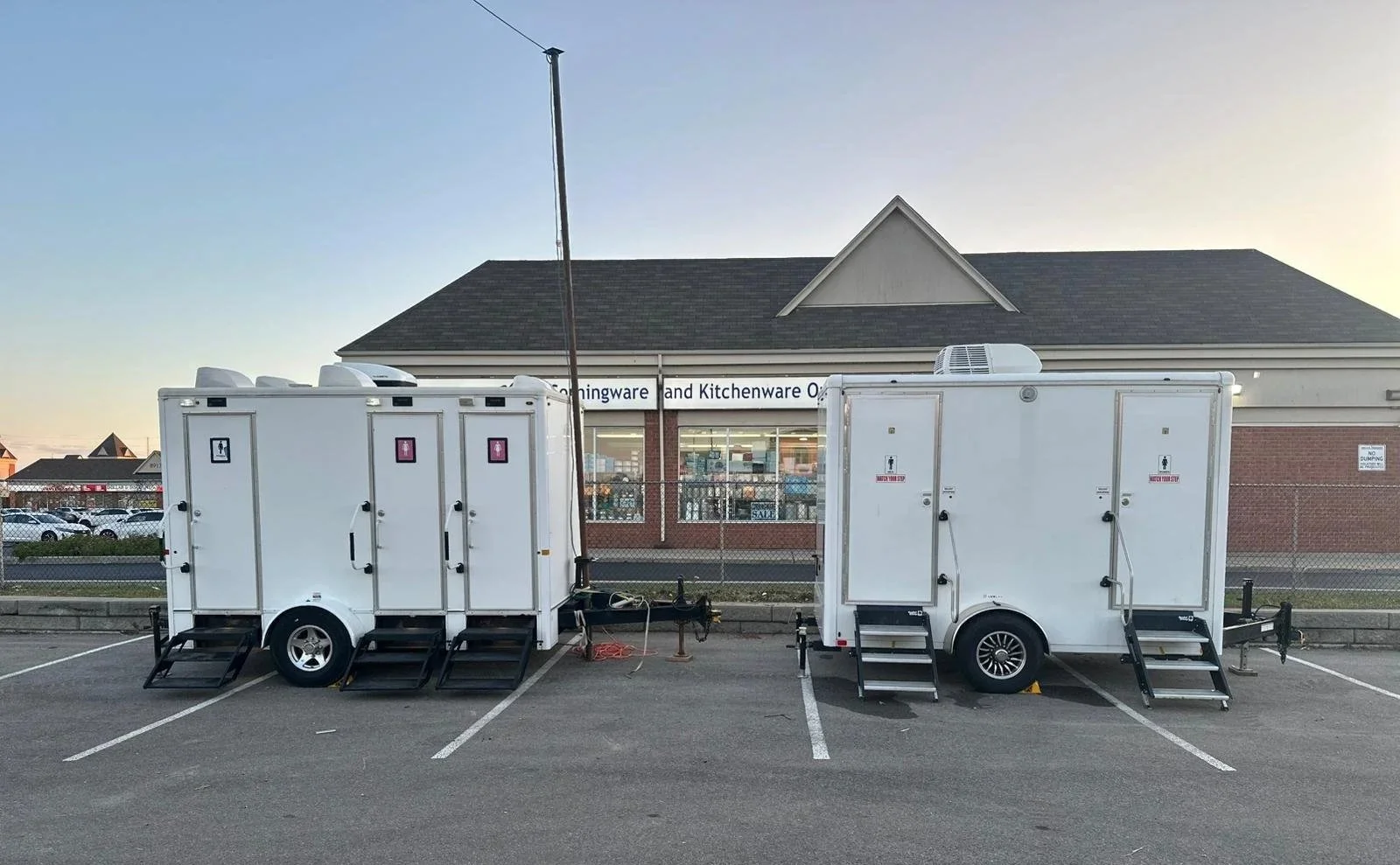 Two white portable restroom trailers parked in a parking lot in front of a building with a sign that reads 'kitchenware and kitchenware o'.