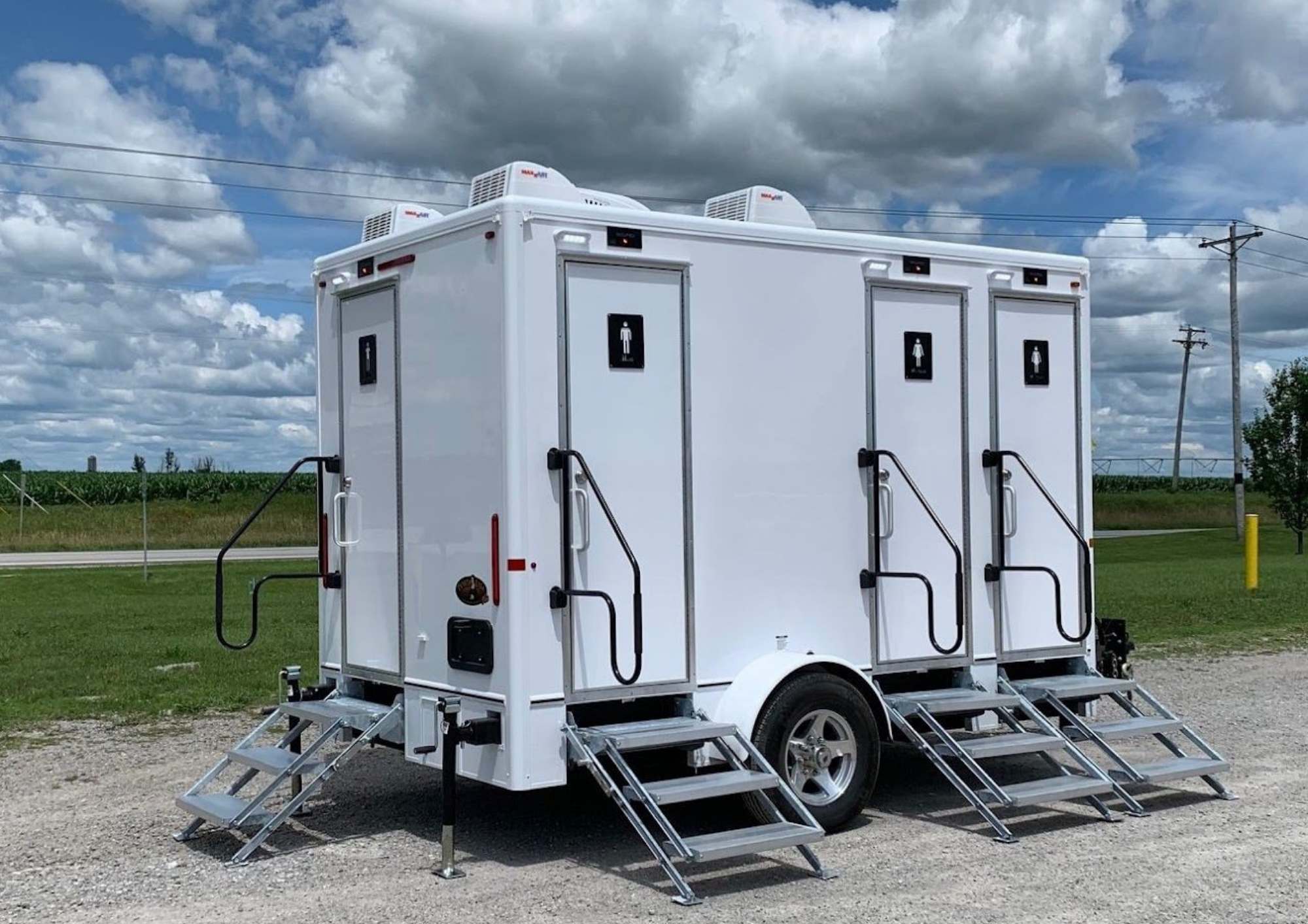 A white four-unit washroom trailer, each with black gender signs and mobile stairs, on a gravel surface outdoors under a cloudy sky.