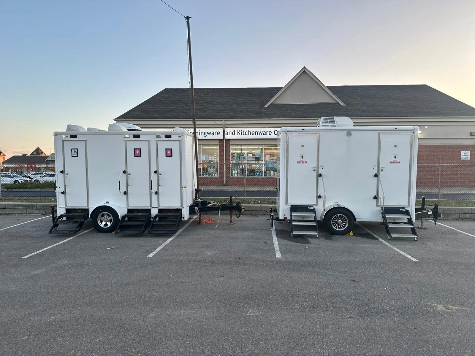 Two white trailer washroom units parked in a lot in front of a building