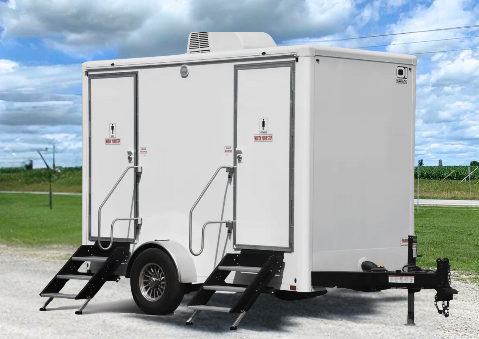 A white portable restroom trailer with two separate stalls, one for men and one for women, situated on a gravel surface with a green field and cloudy sky in the background.