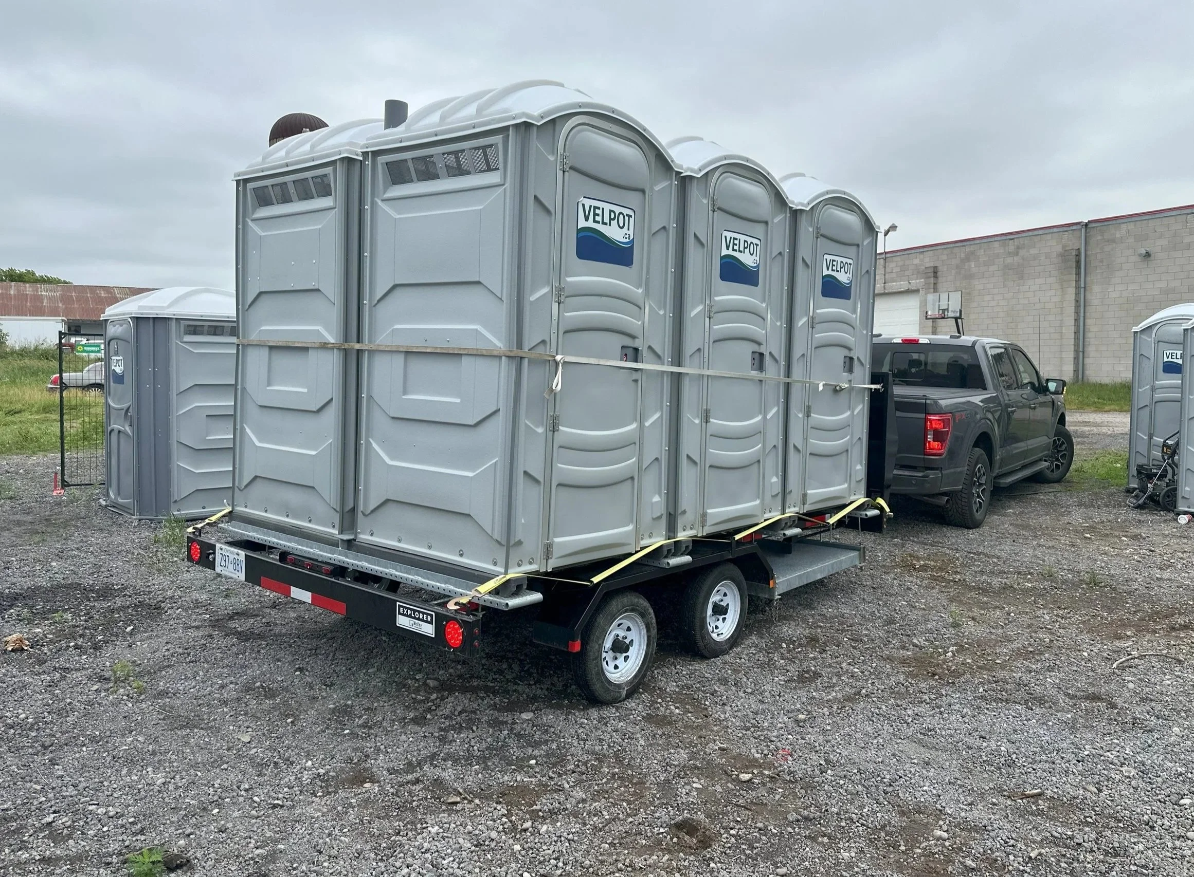 A trailer carrying multiple portable toilets attached to a black pickup truck parked on a gravel lot.