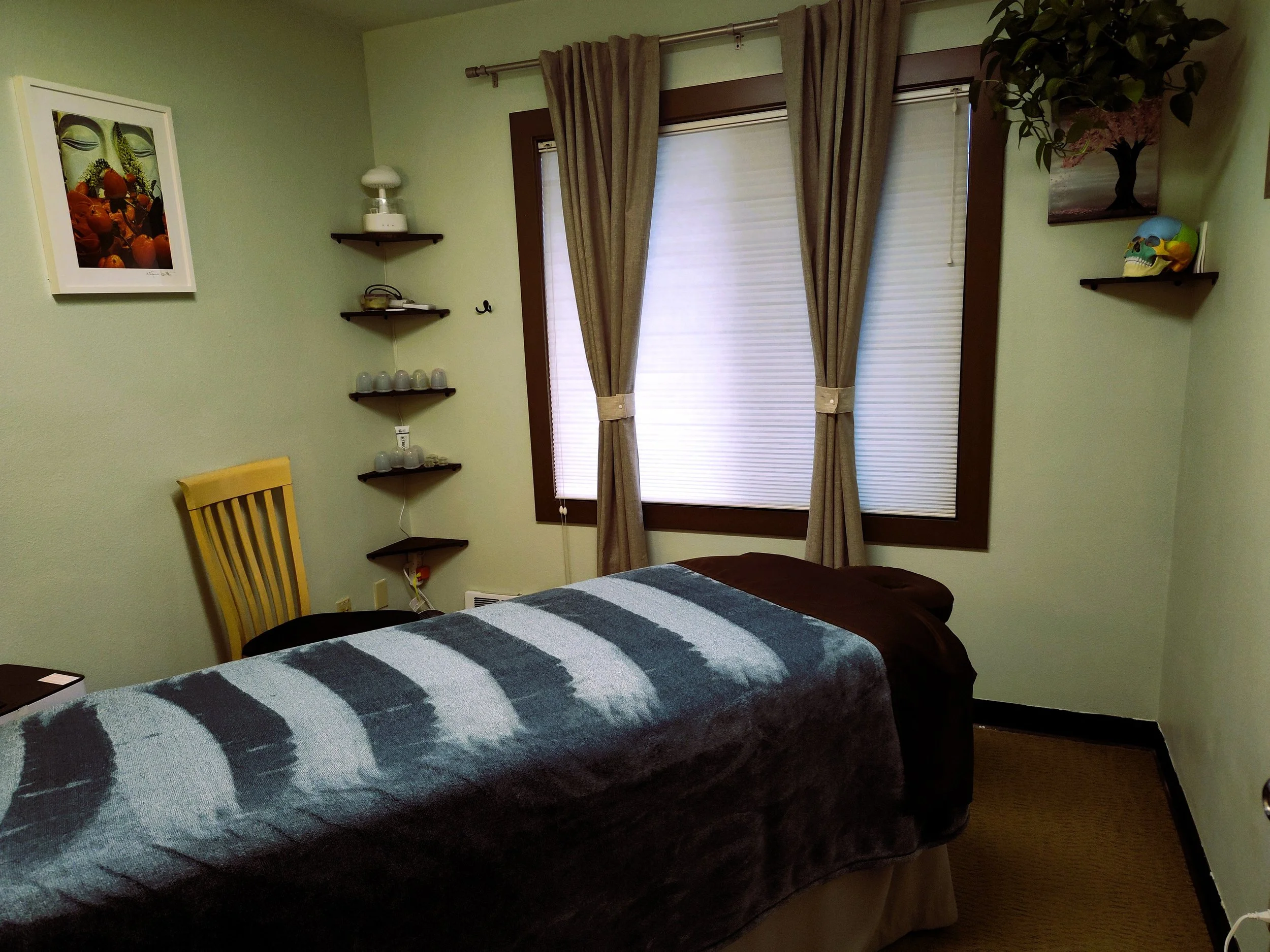A small room with light green walls, a bed with a patterned gray and black comforter, beige curtains on a window with closed blinds, a yellow wooden chair, and wall shelves holding decorative items and a skull.