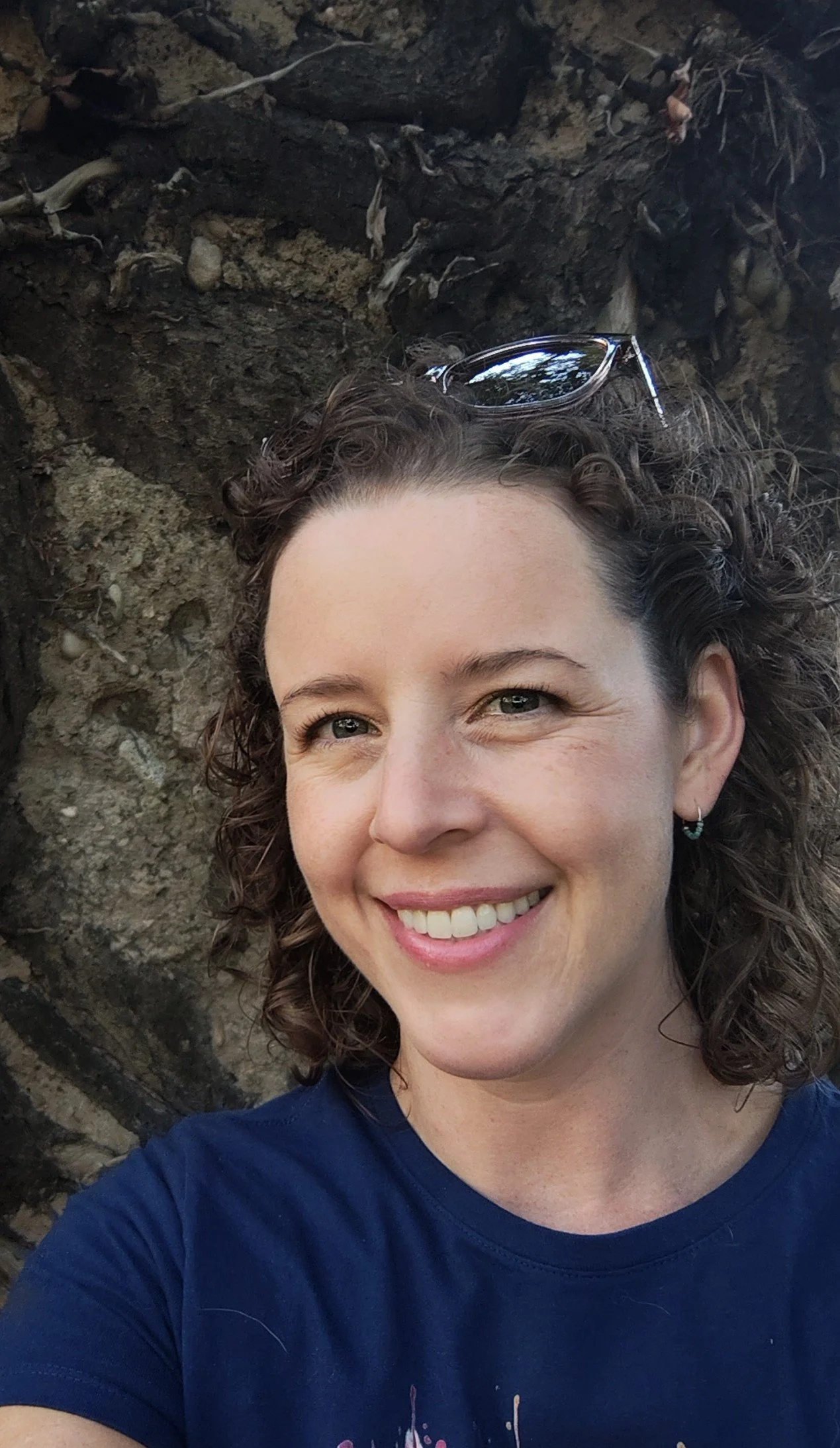 A woman with curly brown hair smiling outdoors, wearing a blue shirt and sunglasses on her head, against a rocky background.
