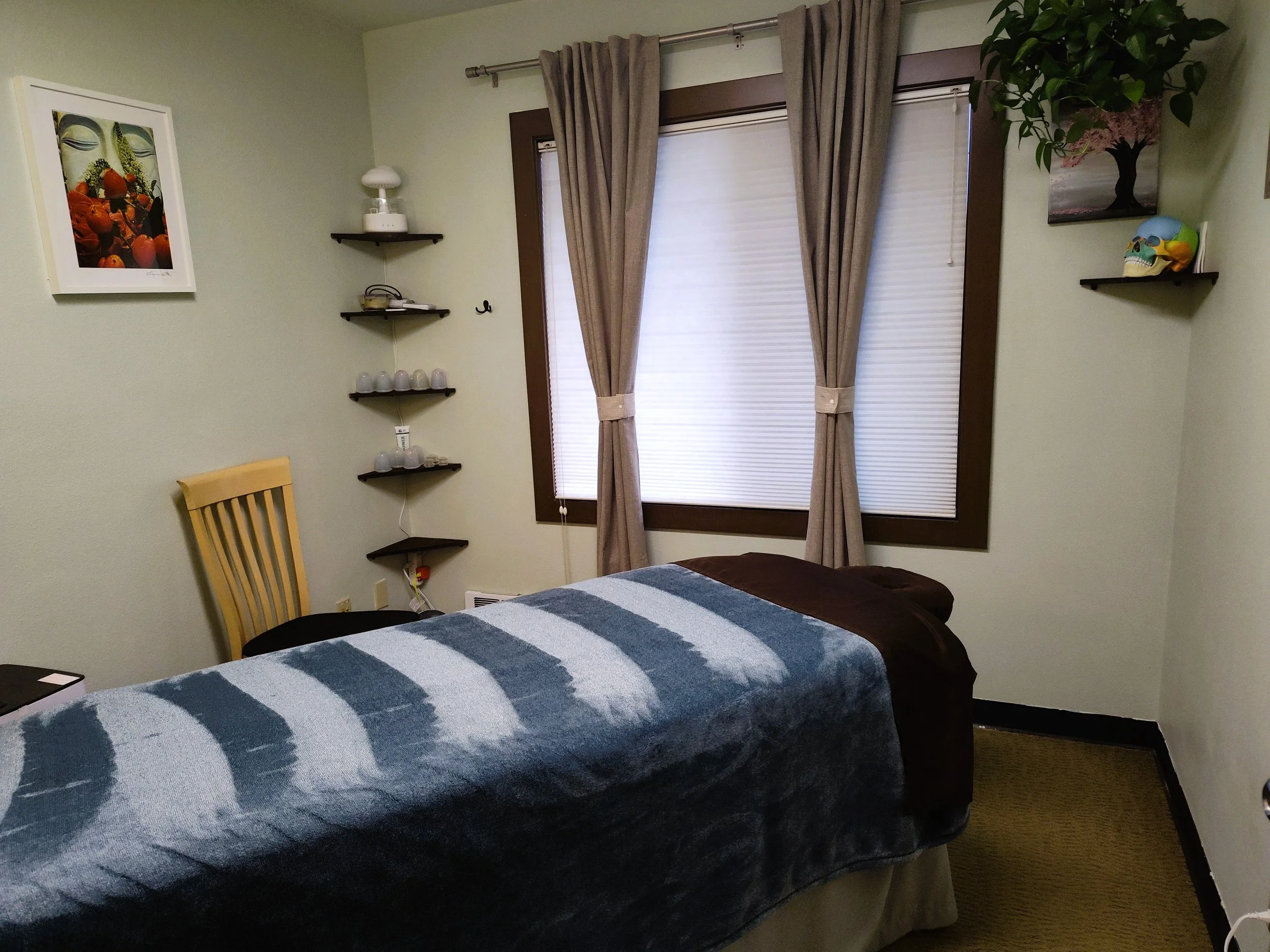 A massage or therapy room with a massage table covered with a blue and gray striped blanket, a yellow wooden chair, and shelves on the wall holding various decorative items, including a colorful picture, a potted plant, and a skull model, with closed beige curtains over a window.