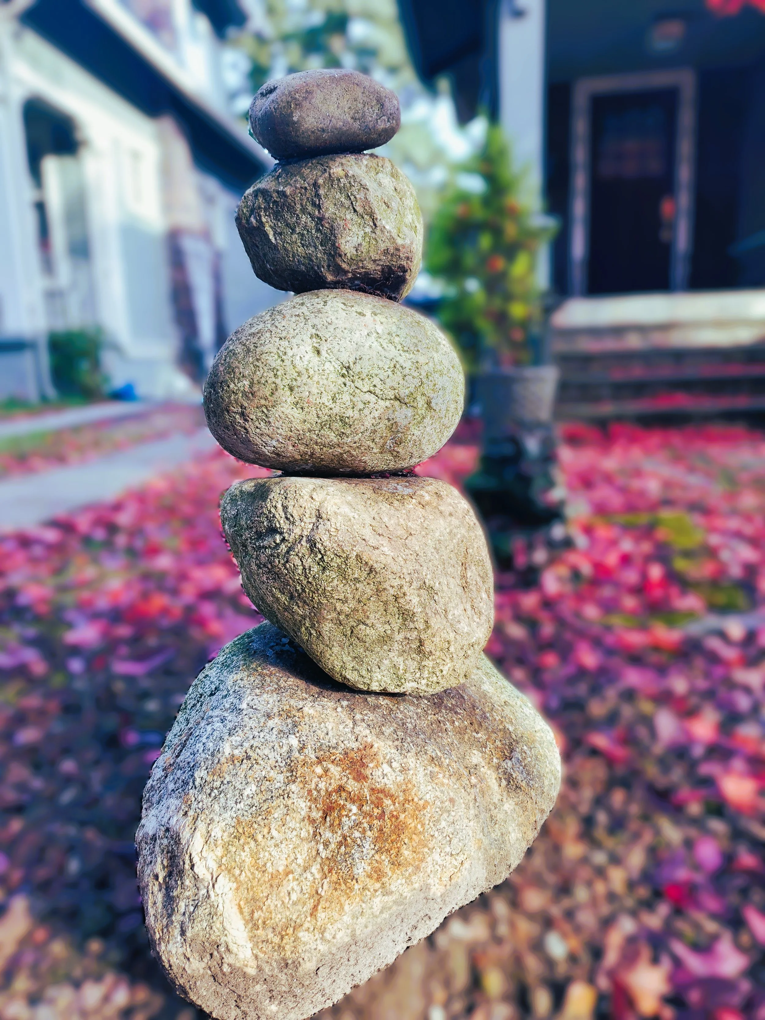 Stack of five smooth stones balanced on top of each other in front of a house with colorful fall leaves on the ground.