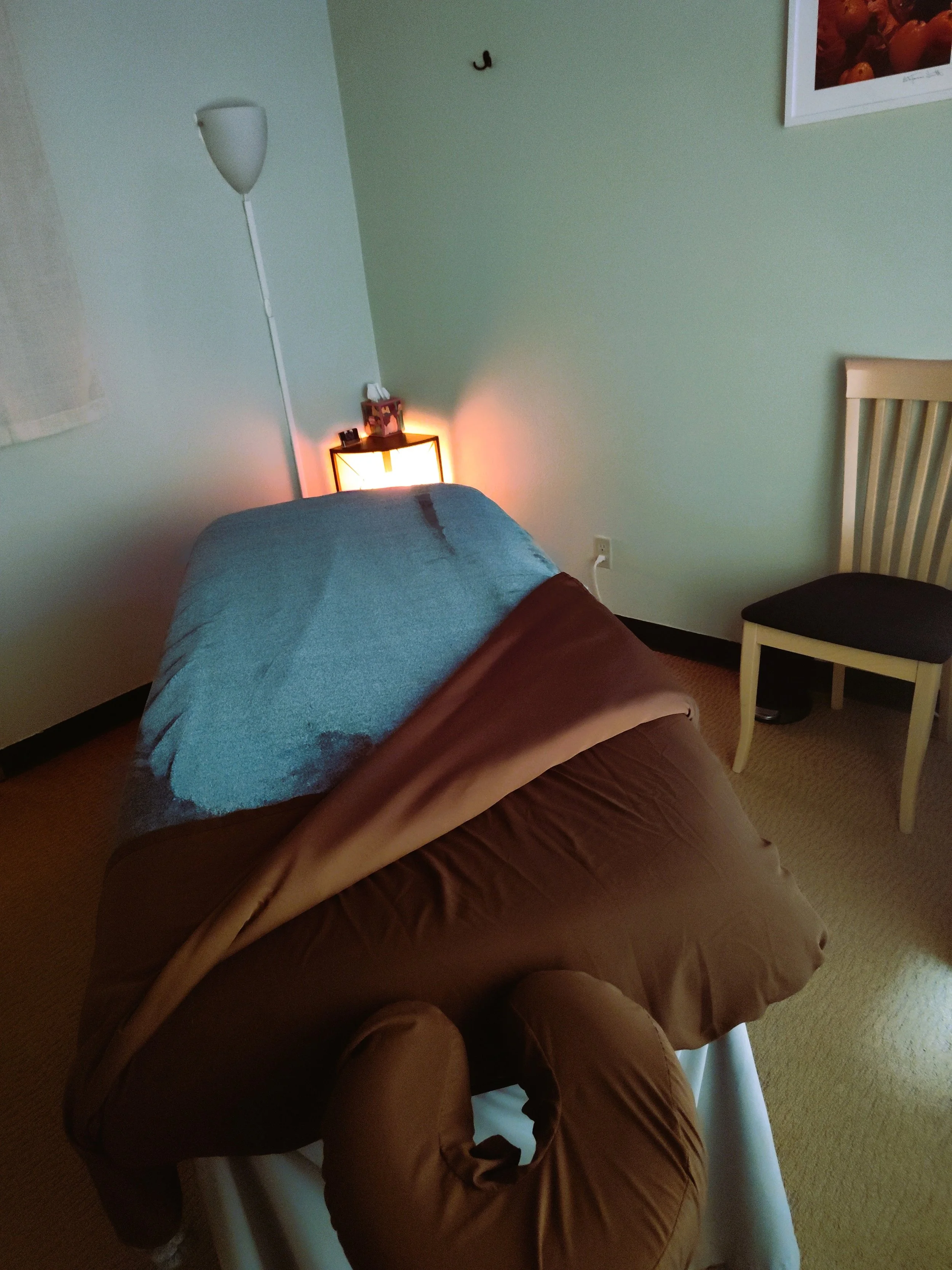 Massage table with brown and blue blankets in a room with a green wall, a white lamp, a small side table with a light, a framed picture, and a wooden chair.