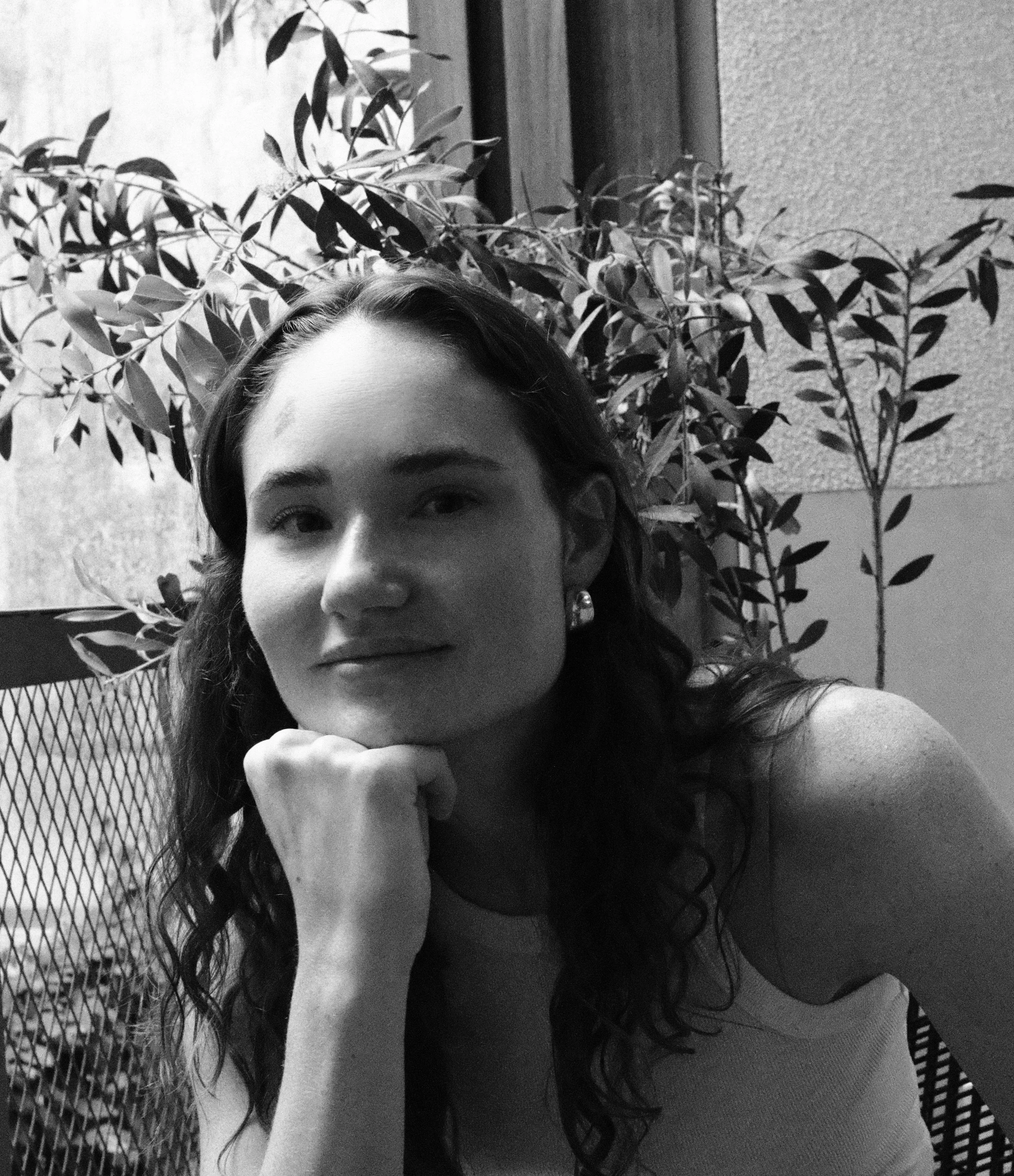 A young woman with wavy hair and earrings resting her chin on her hand, sitting in front of a leafy plant and a textured wall, in black and white.