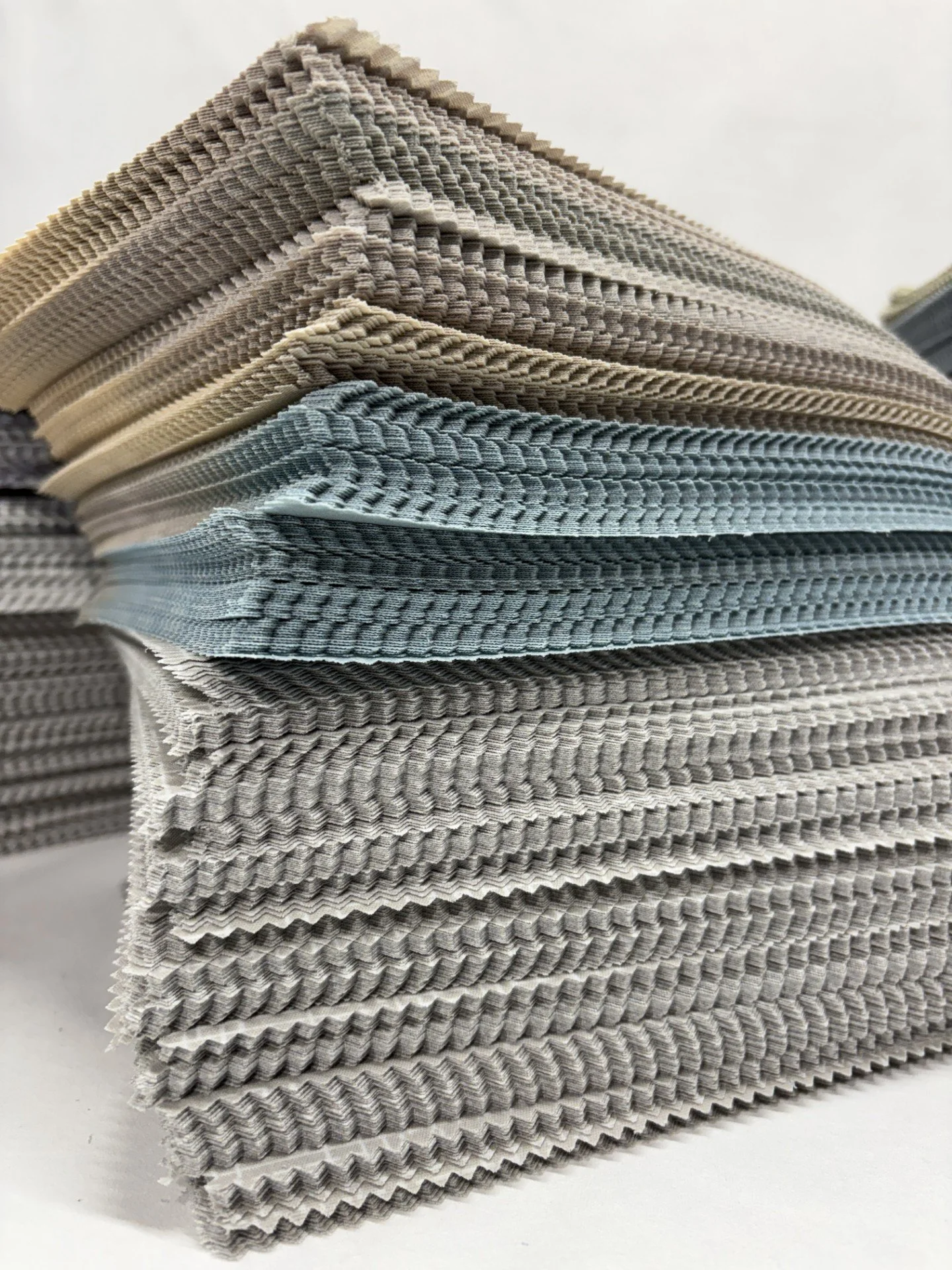 Stacked fabric swatches in various colors and textures, including beige, gray, and blue, with textured and woven patterns.