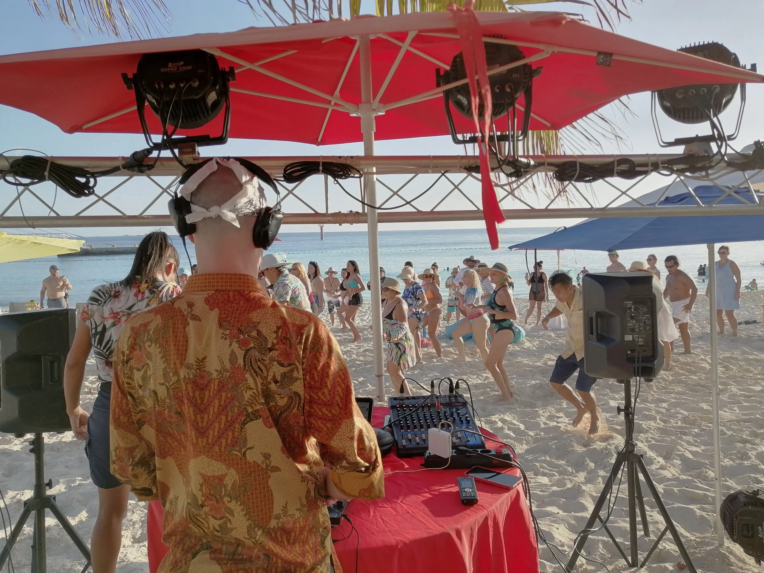 People dancing on the beach during a DJ event under a red canopy, with a person in the foreground wearing a patterned shirt and headphones, and DJ equipment on a table.