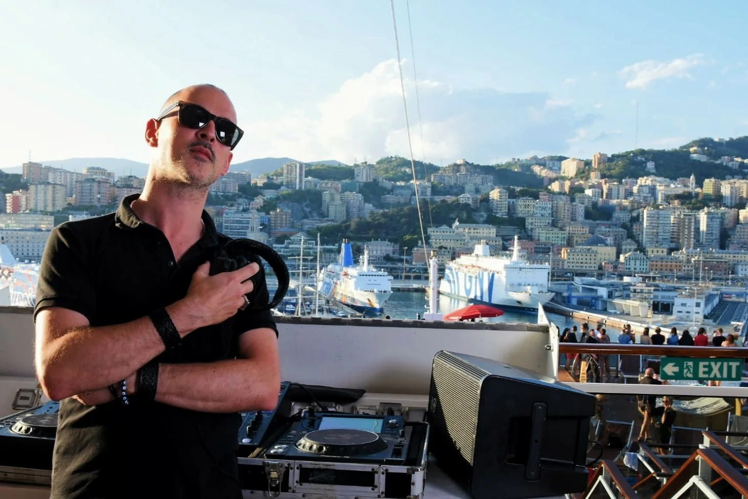 DJ with headphones standing at outdoor event with a marina and hillside cityscape in the background.