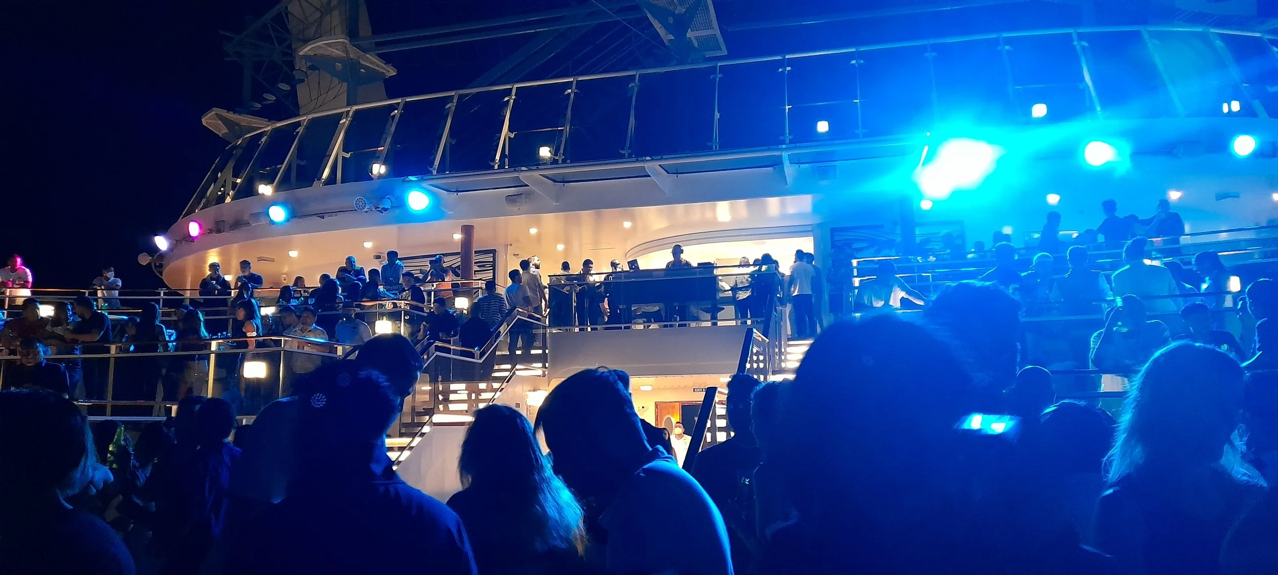 People gathered on a cruise ship deck at night, illuminated by blue and purple lights, with multiple levels and railings visible.
