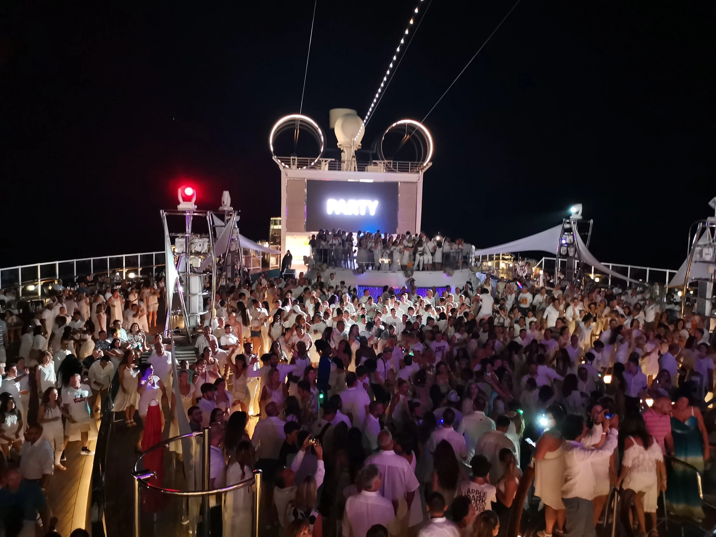Crowded deck of a cruise ship at night, with a large group of people dancing and socializing near a DJ booth that displays the word 'PARTY'. The ship's structure, ropes, and lighting equipment are visible, with a dark sky above.