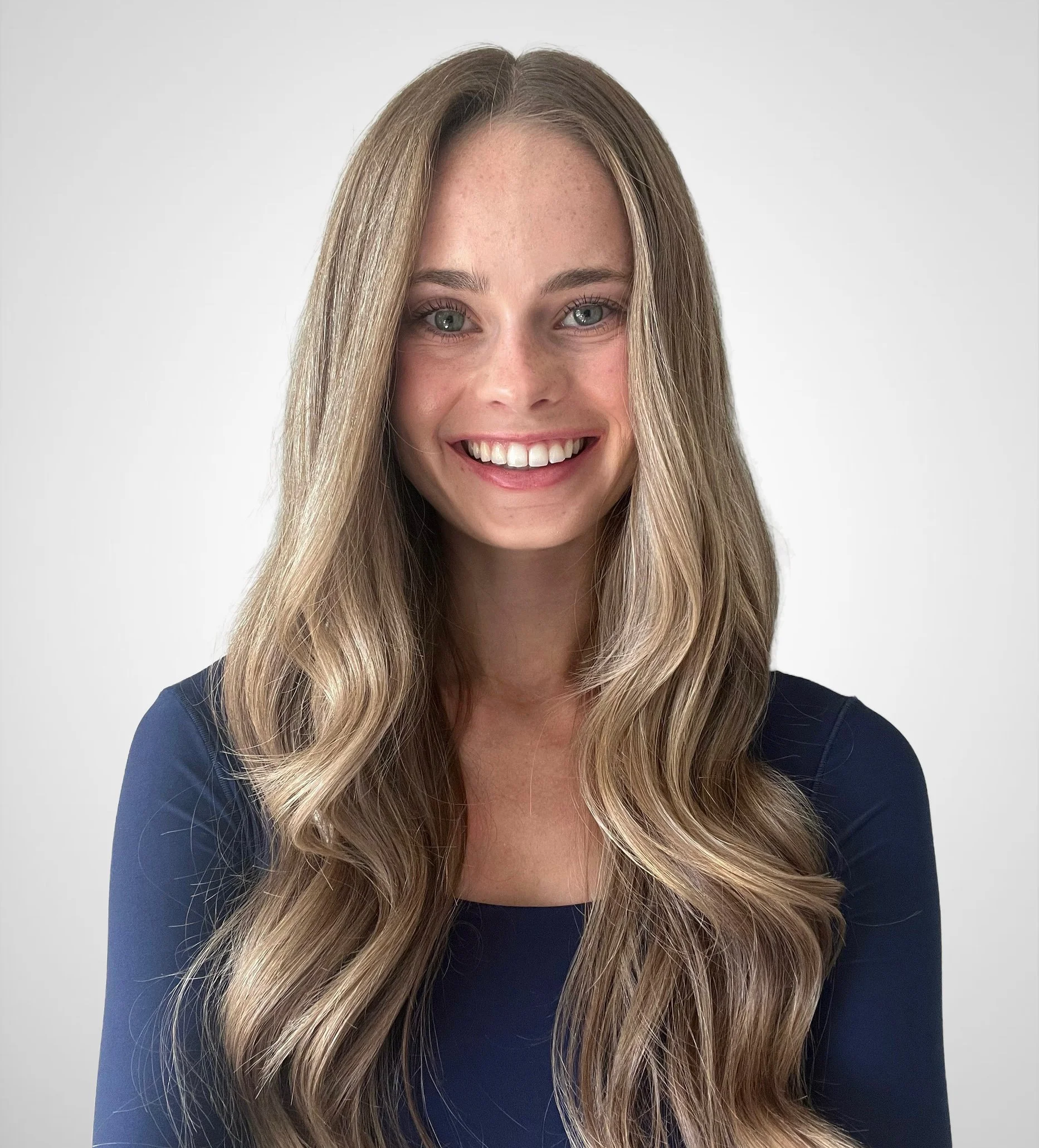 A woman with long wavy blonde hair, bright blue eyes, and a big smile, wearing a navy blue top, against a plain white background.