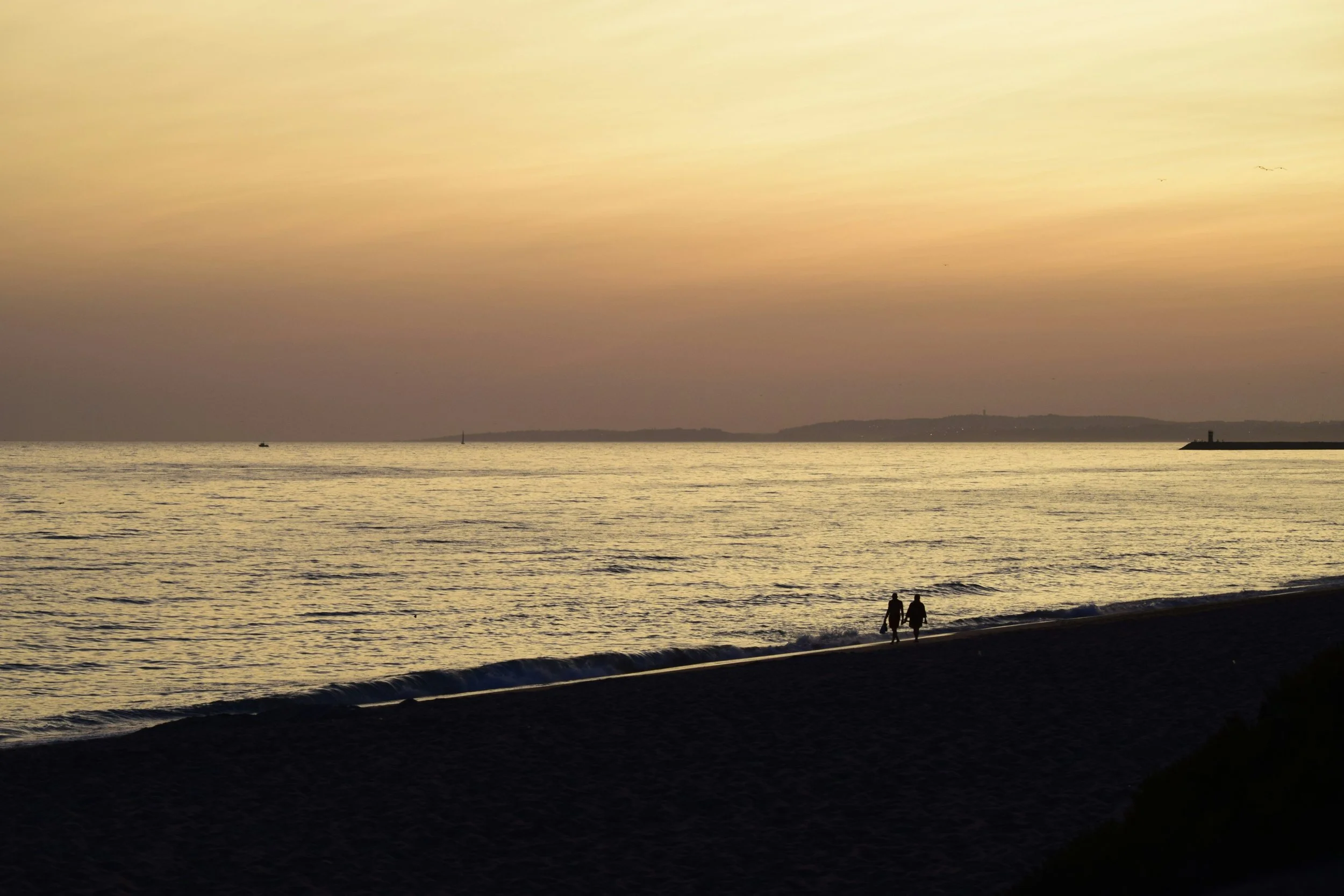 A couple walks along the shoreline at sunset, with calm ocean waters, distant land, and a colorful sky.