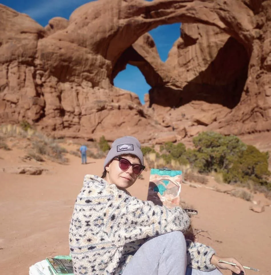 A woman painting a landscape on a canvas in a desert with large rock formations and natural arches in the background.