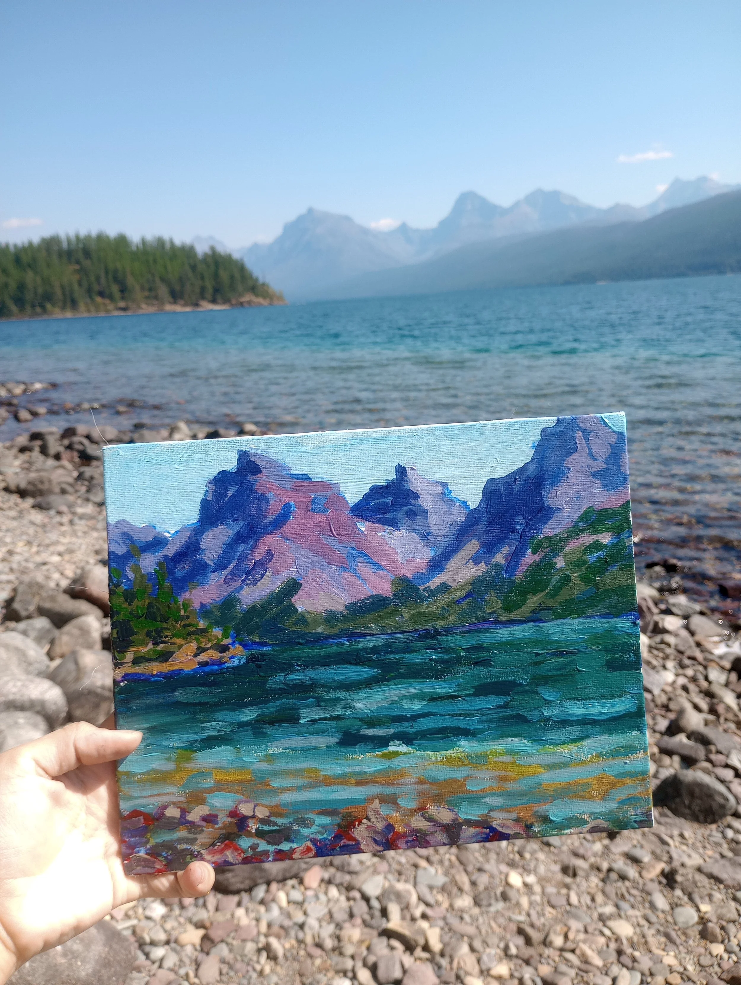 A person holding a small painting of a mountain landscape near a lake, with rocky shores and a forest, against a larger lake and mountain range backdrop.