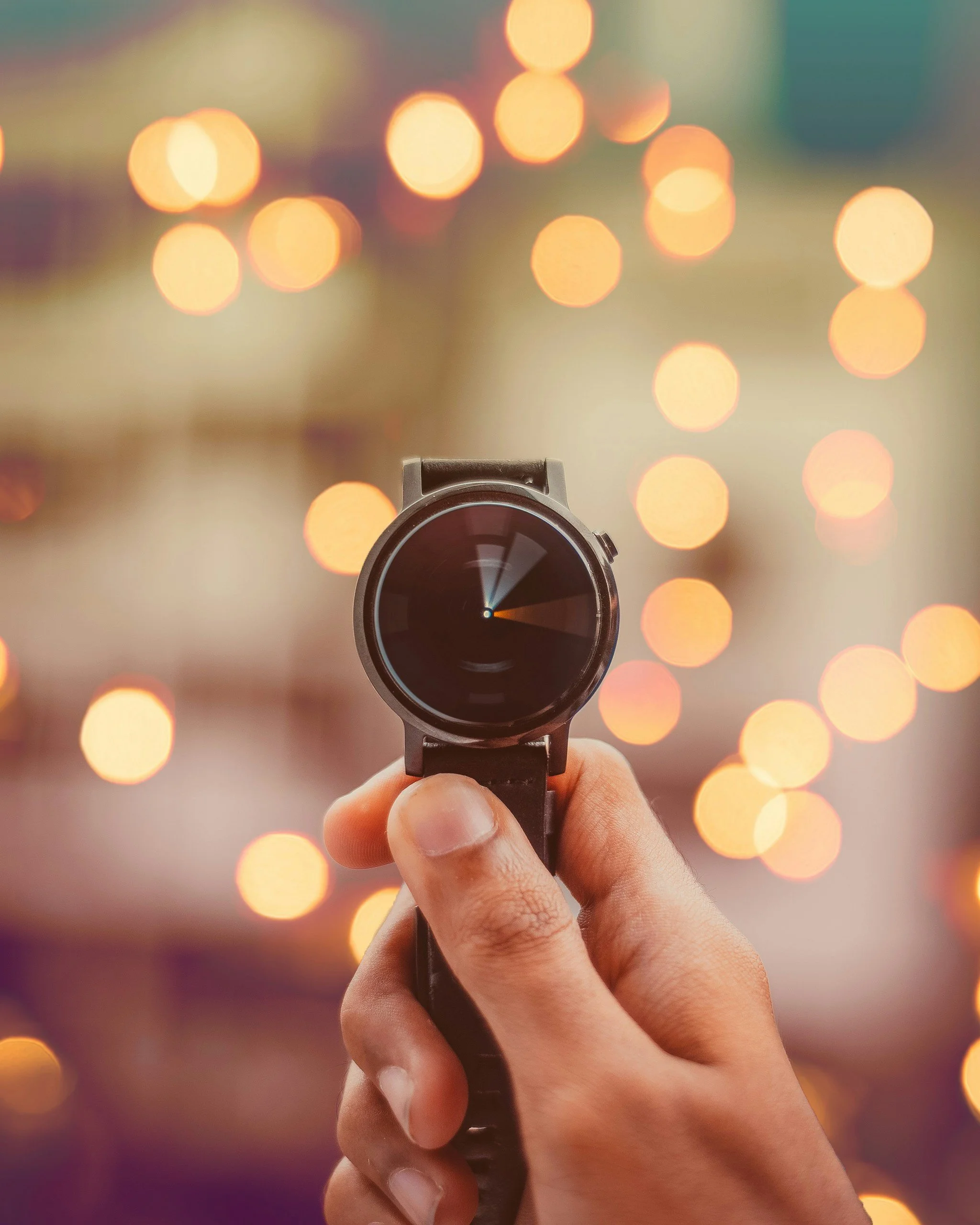 Una mano sostiene un reloj inteligente negro en primer plano, con un fondo de luces difusas de colores cálidos.