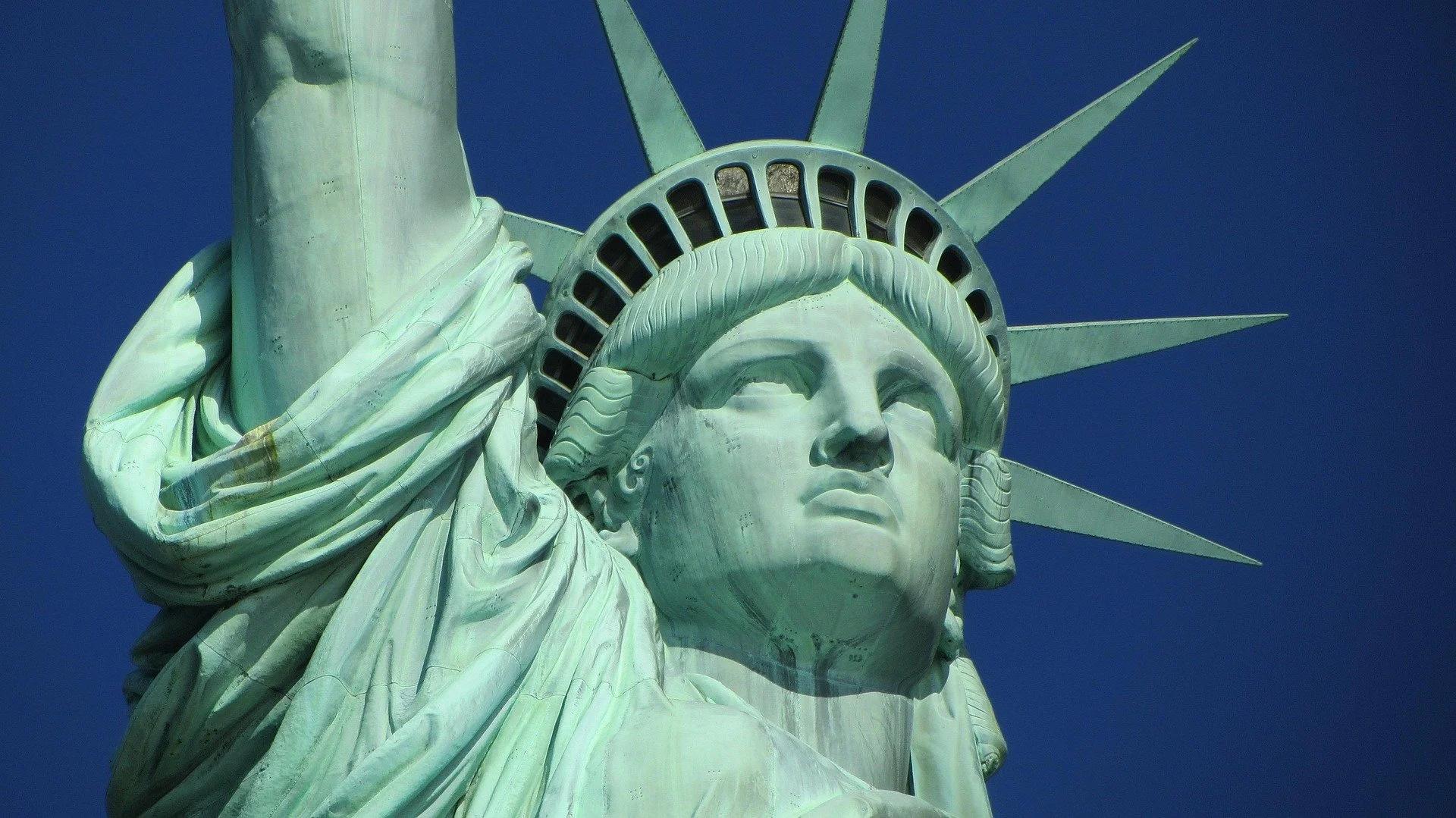 Primer plano de la Estatua de la Libertad, destacando su rostro y la corona con picos, contra un cielo azul.