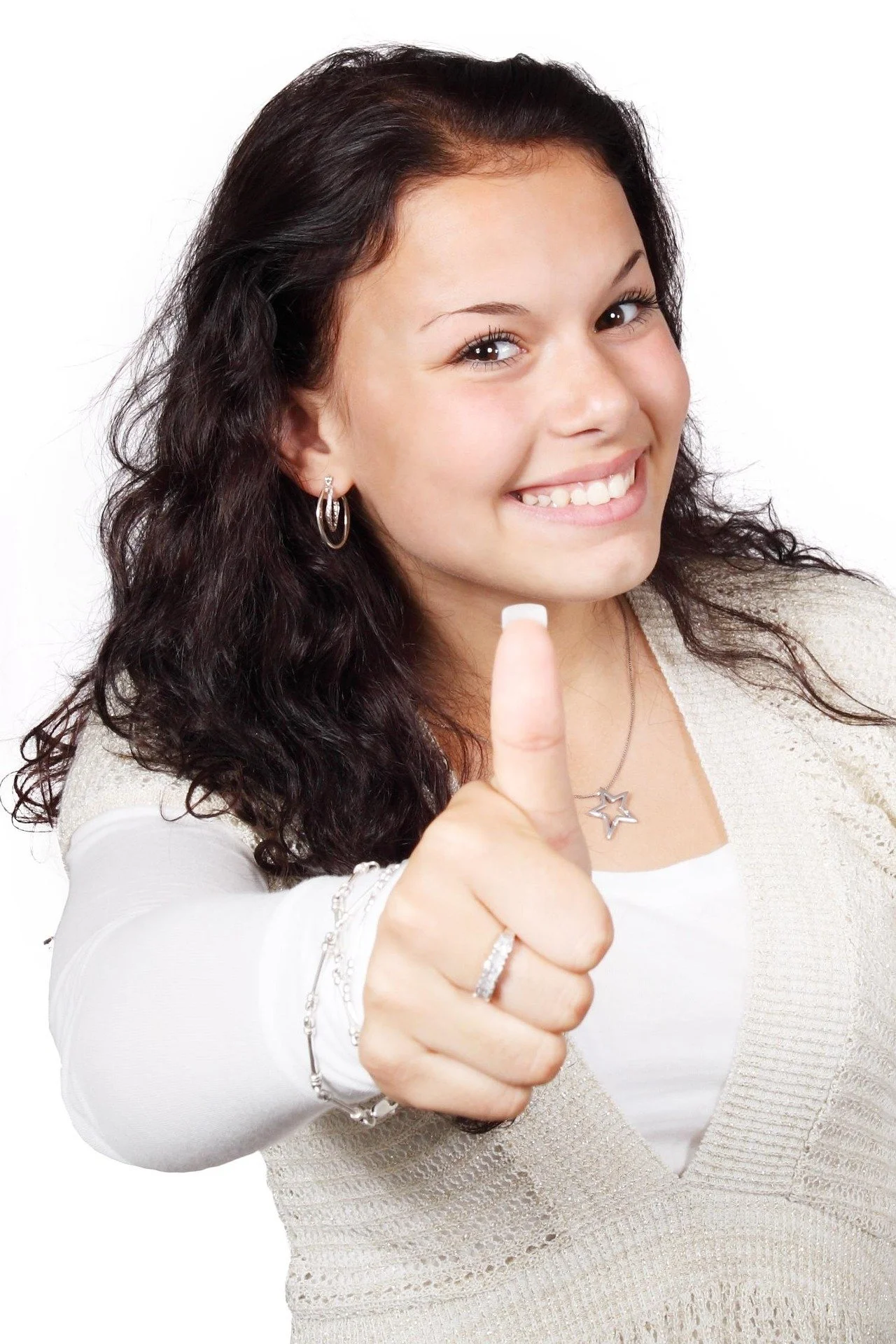 Joven mujer con cabello rizado oscuro, sonriendo, mostrando el pulgar hacia arriba, con aretes de aro y un collar en forma de estrella, vestida con una blusa blanca y un suéter beige, fondo blanco.
