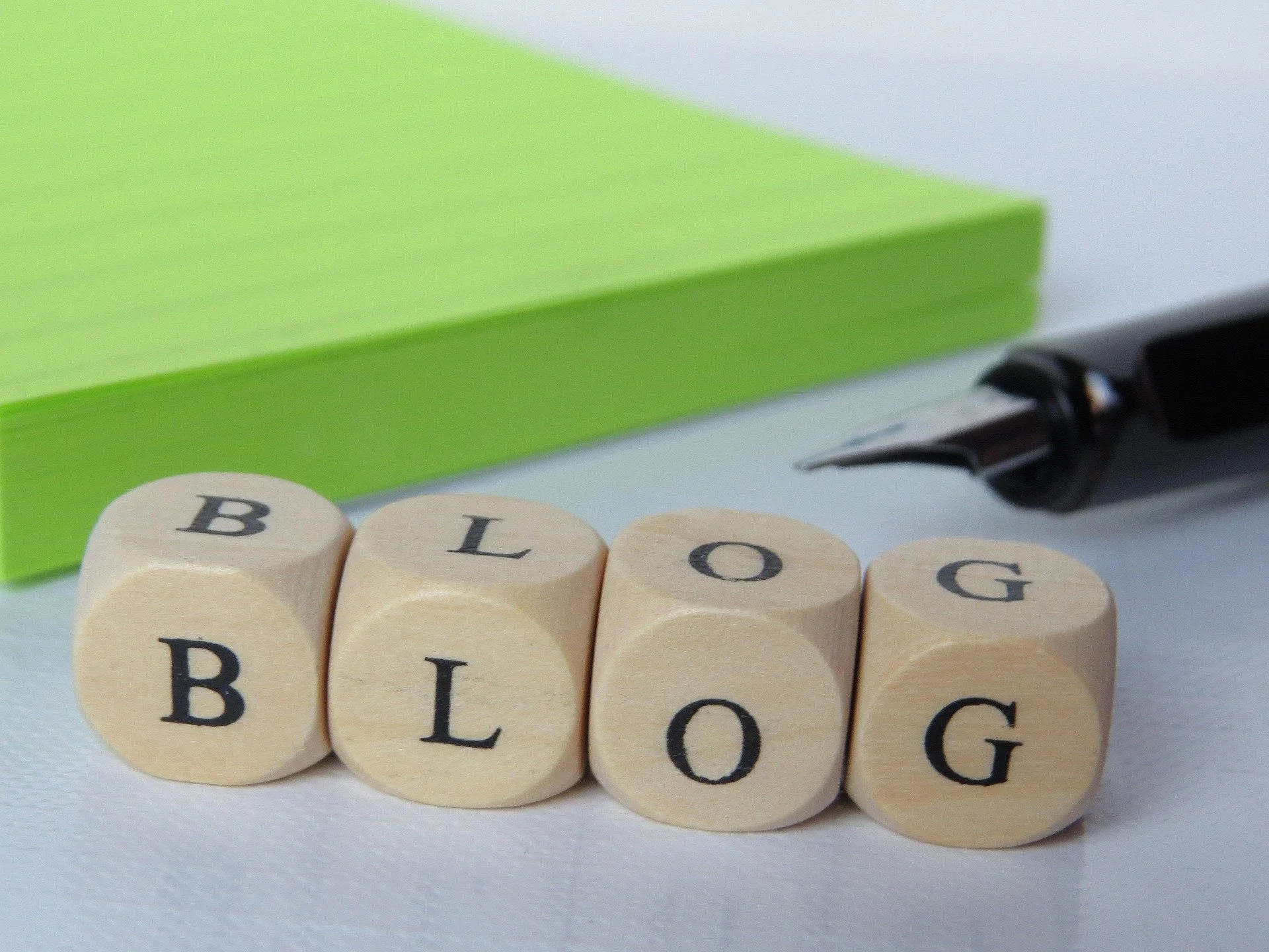 Cuatro bloques de madera con letras que forman la palabra 'BLOG', una caja verde, y un bolígrafo con tapa negra en un fondo blanco.