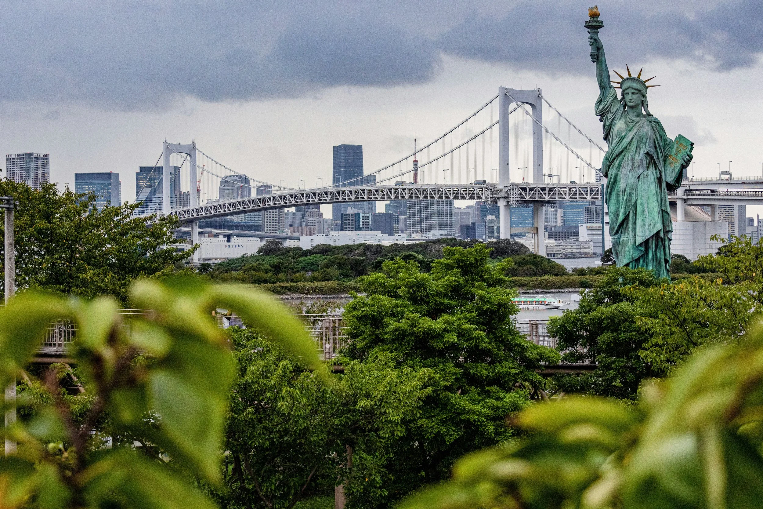 Reproducción de la Estatua de la Libertad en un parque con árboles y un puente en el fondo, en una ciudad con edificios modernos y cielo nublado.