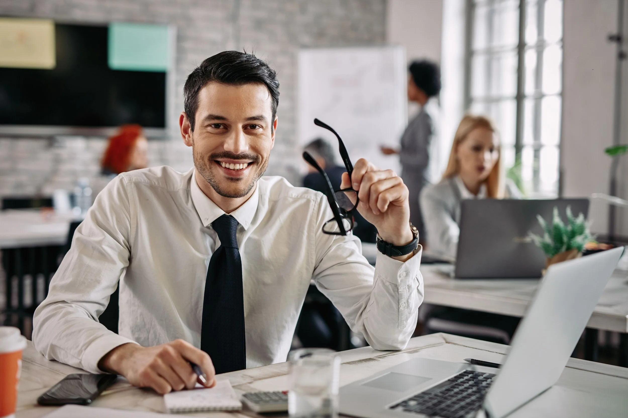 Hombre de negocios sonriente en una oficina moderna, sosteniendo gafas de sol, con laptop, cuaderno y teléfono en el escritorio, en un ambiente de trabajo con otras personas en el fondo.