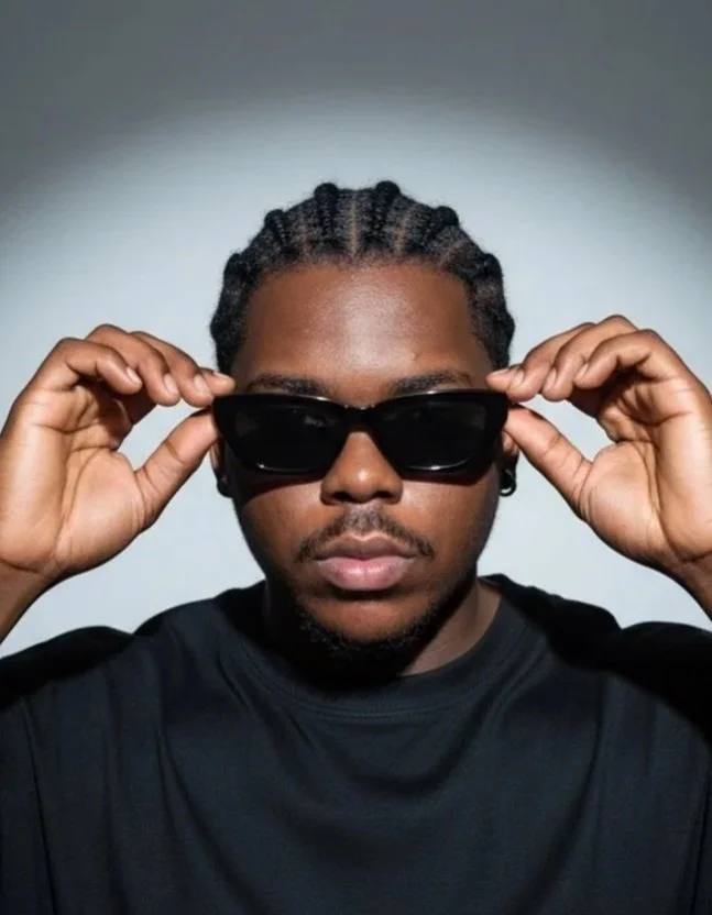 A young man with braided hair wearing black sunglasses and a black shirt, holding the sunglasses with both hands in front of a plain background.