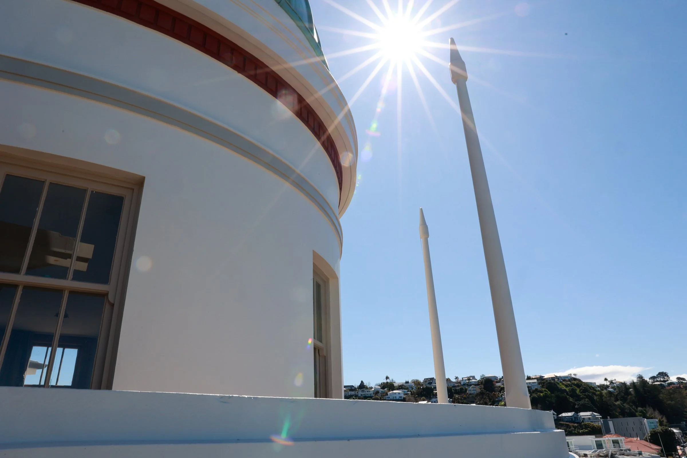 Close-up of a white building with curved architecture, windows, and flagpoles, under a bright sun in a clear blue sky.