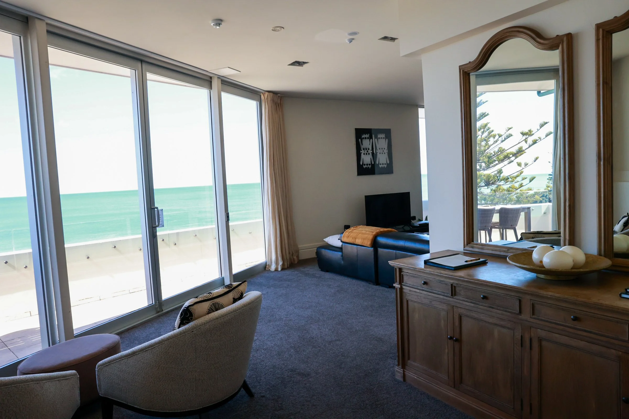 Living room with large sliding glass doors leading to a balcony, ocean view, beige armchair, wooden dresser with mirror, flat-screen TV, black leather sofa with yellow blanket, decorative vase with white eggs, and a balcony with outdoor chairs and ta