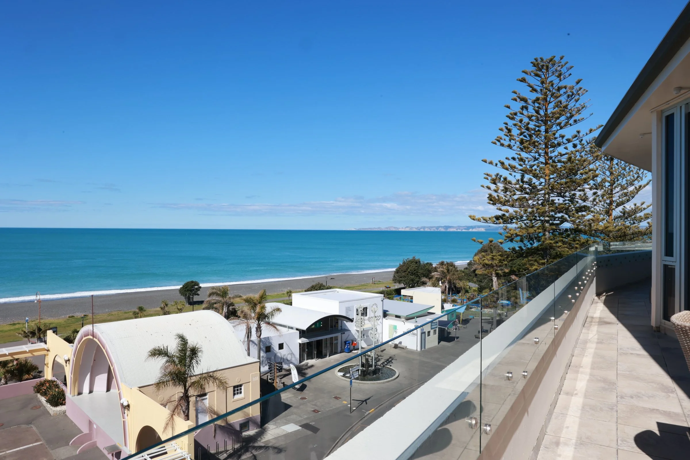 Ocean view from a balcony overlooking a beach and small buildings, with a large pine tree to the right and clear blue sky.