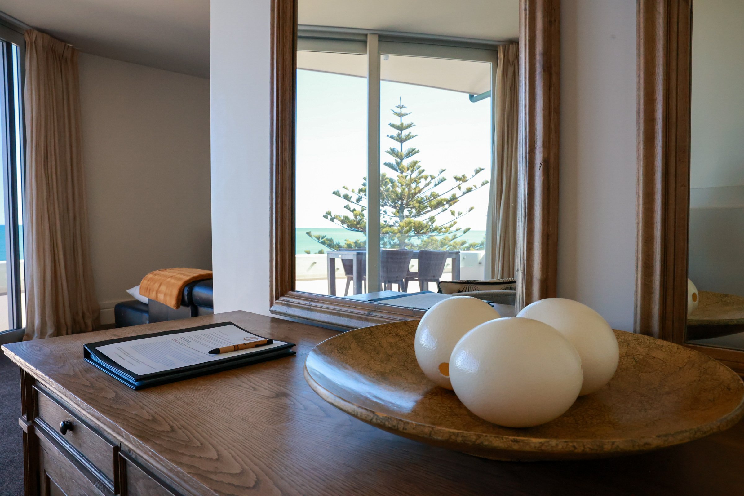 Interior of a room with a wooden desk, a large mirror, and a view of a balcony outside featuring a chair, table, pine tree, and ocean in the background.