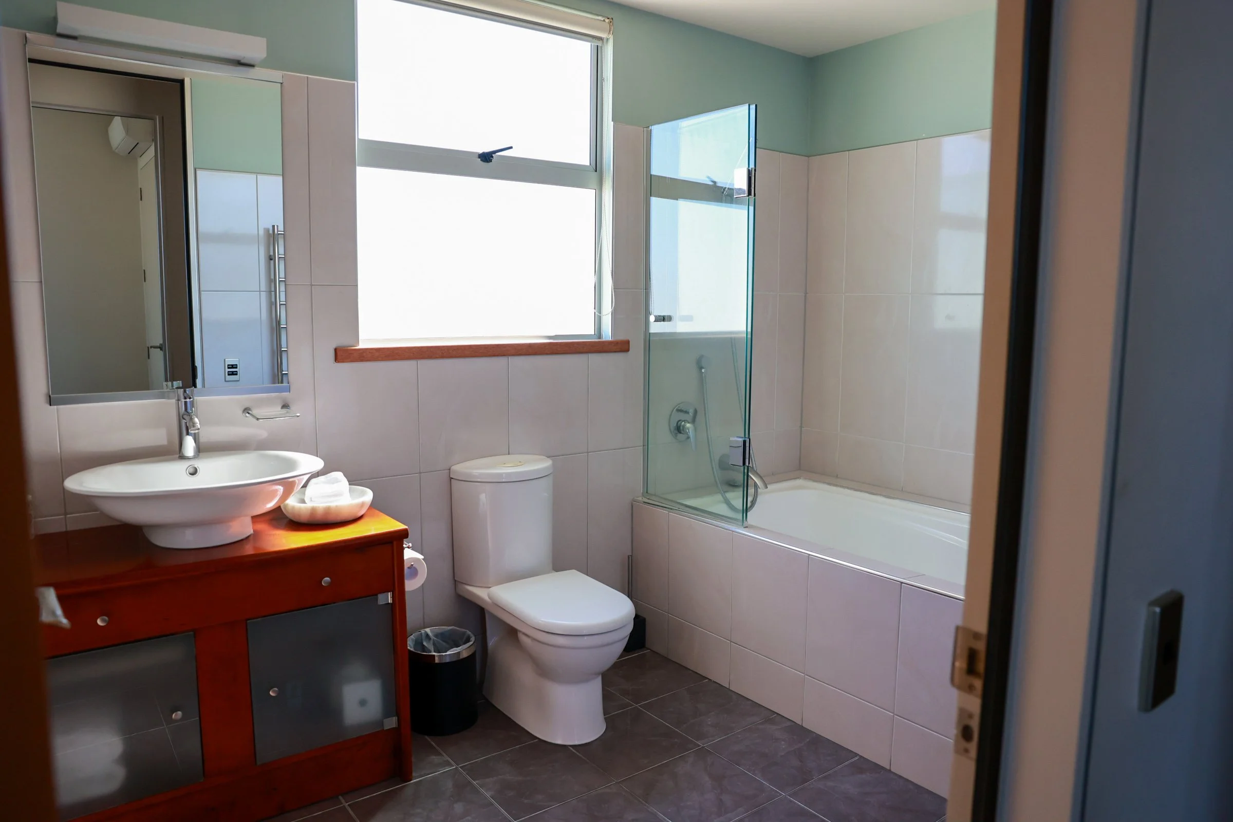 Bathroom with a white toilet, a wooden vanity with a vessel sink and mirror, a window, and a bathtub with a glass shower panel.