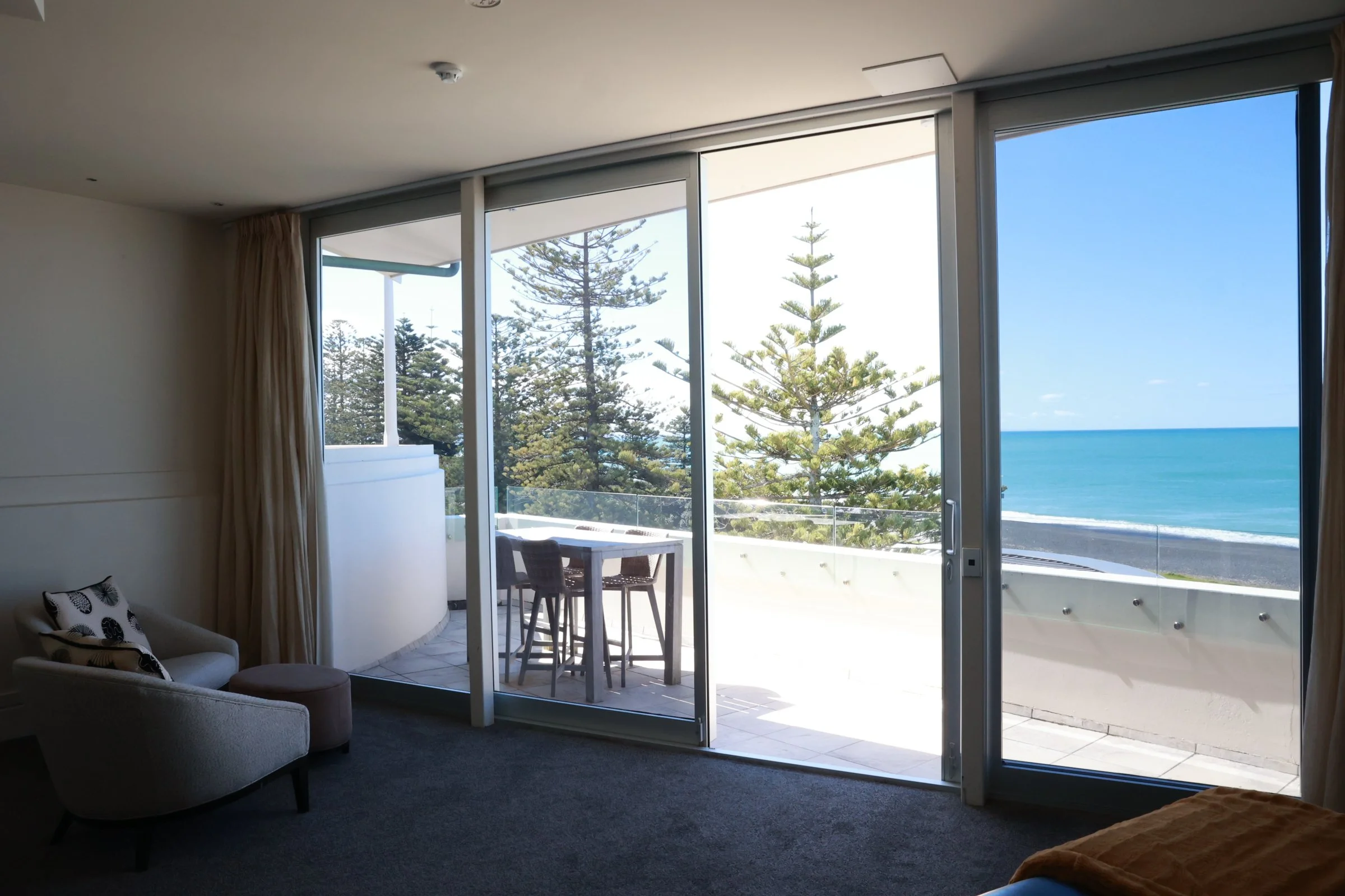 Interior of a room with a glass sliding door leading to a balcony with outdoor furniture, overlooking pine trees and the ocean.