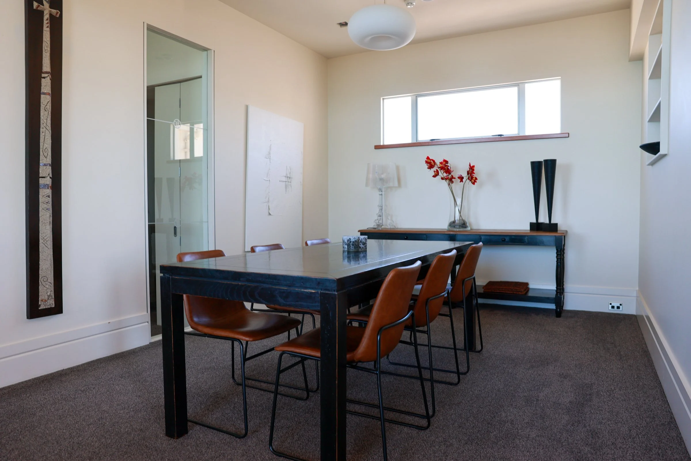 Dining room with a large wooden table, six brown chairs, and a black console table against the wall with a glass vase with red flowers and decorative black objects. Light-colored walls and a small window.