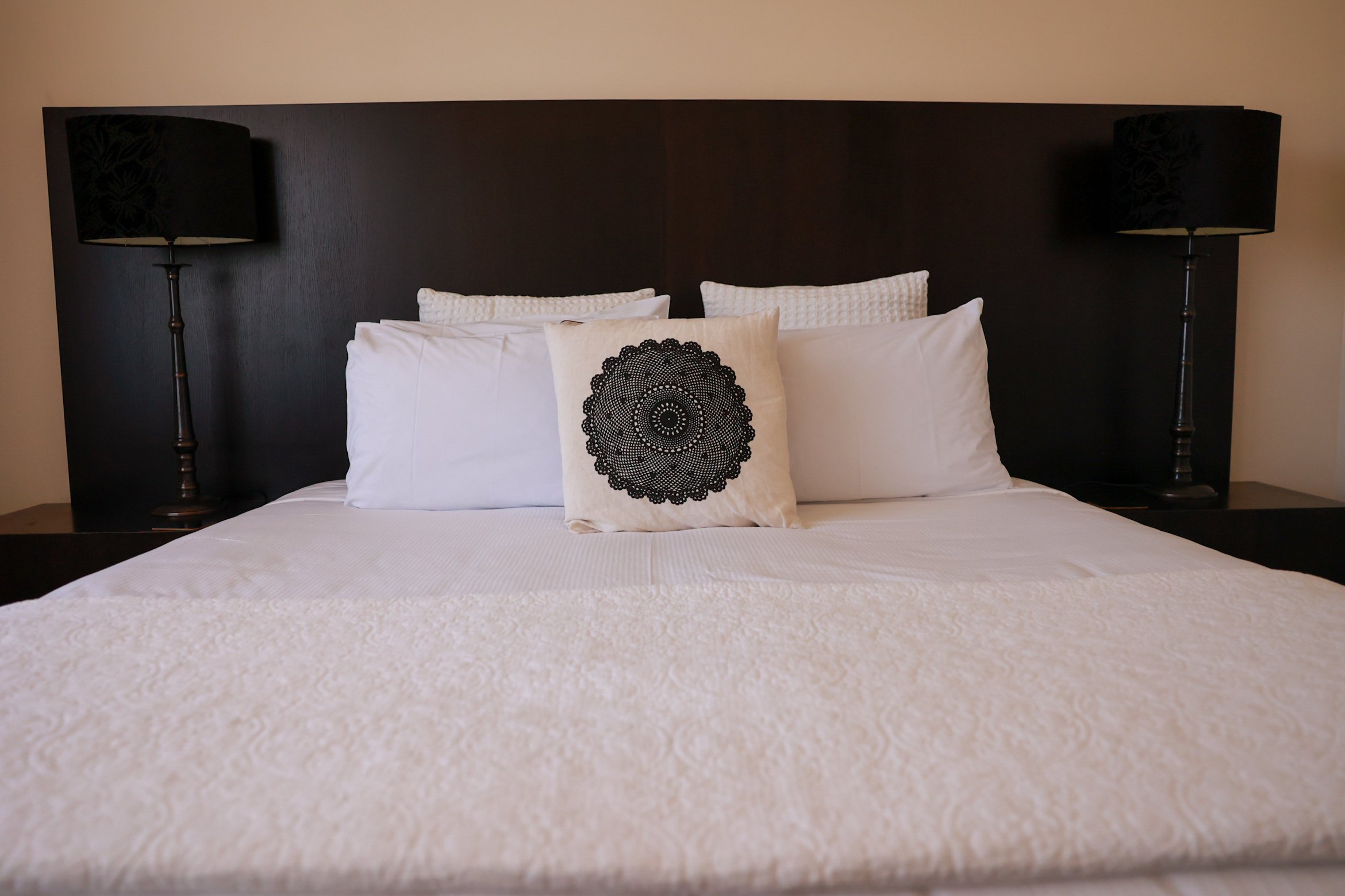 Made bed with white bedspread, multiple white pillows, and a decorative pillow with black lace pattern, black headboard, and black lamps on bedside tables.