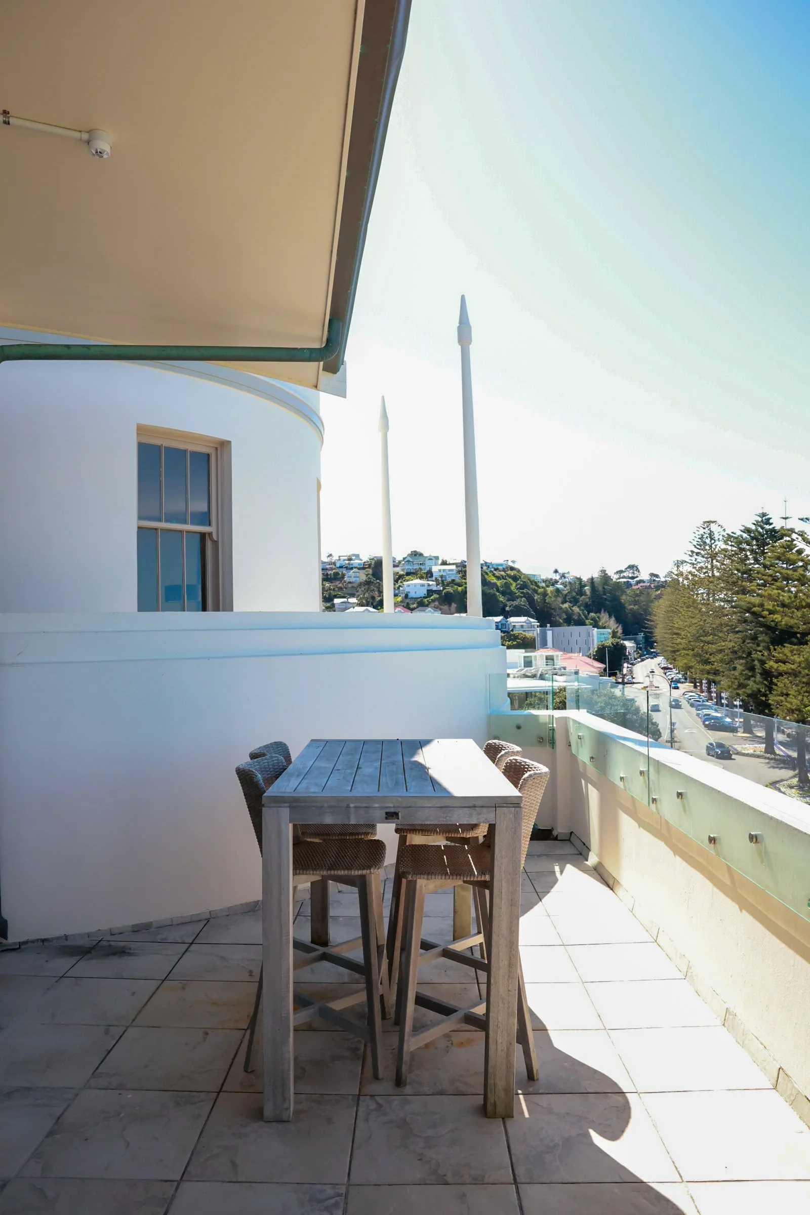 Empty balcony with a wooden table and four chairs, overlooking a street lined with trees and buildings under a clear sky.