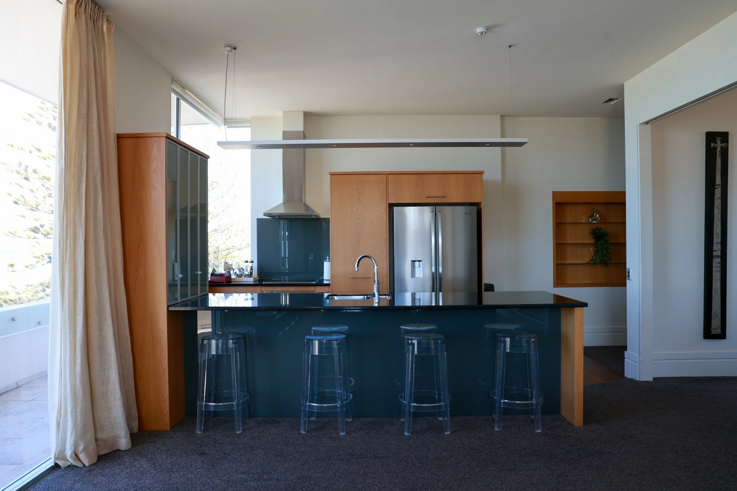 Modern kitchen with a black countertop island, transparent bar stools, stainless steel refrigerator, wooden cabinetry, and large windows with beige curtains.