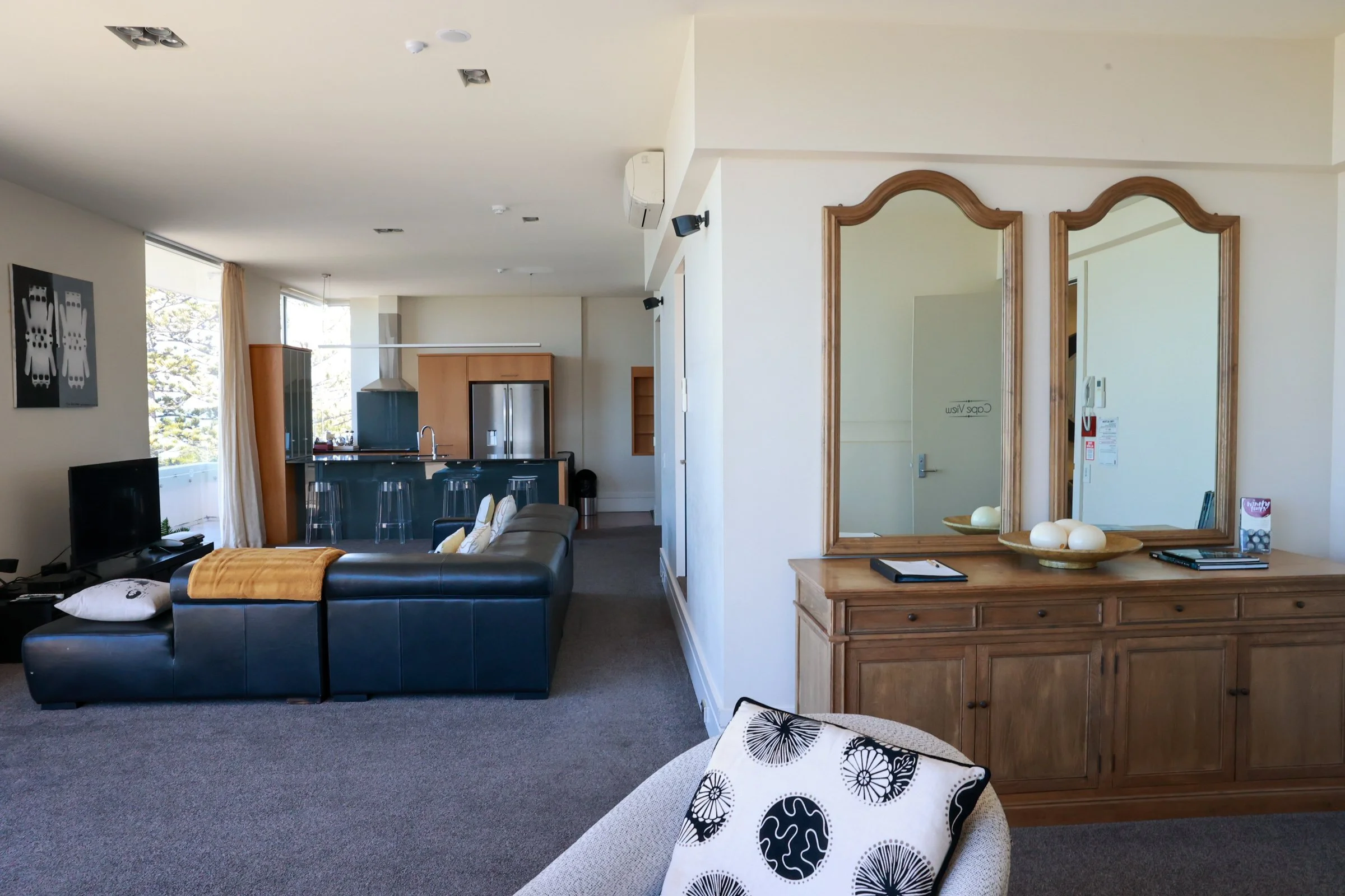 Living room with black leather sectional sofa, television, and kitchen area in the background featuring a refrigerator, countertop, and bar stools; wooden sideboard with two mirrors, decorative bowls, and magazines.