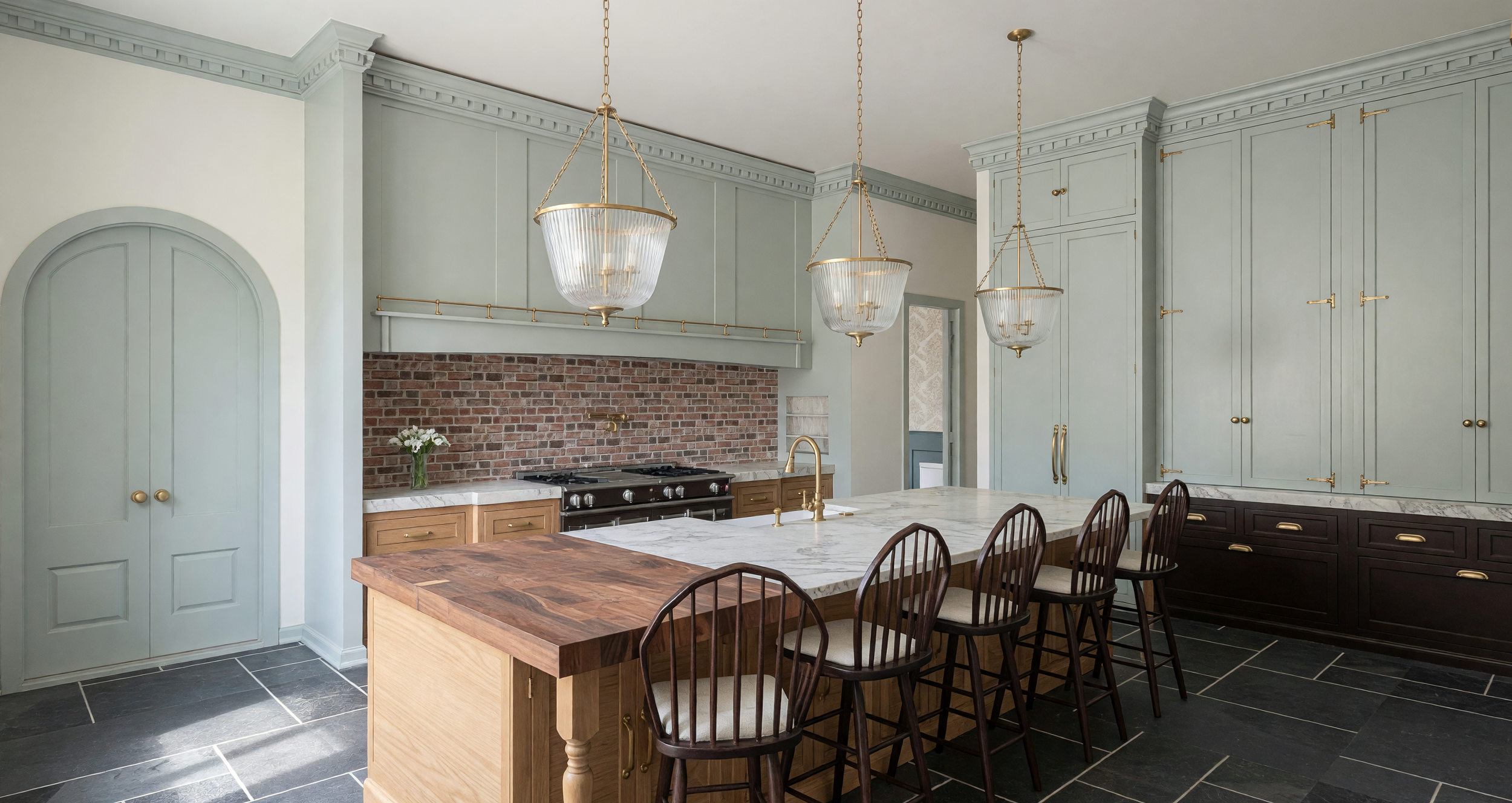 Modern kitchen with light green cabinets, a brick backsplash, a marble countertop island, black chairs, and hanging pendant lights.