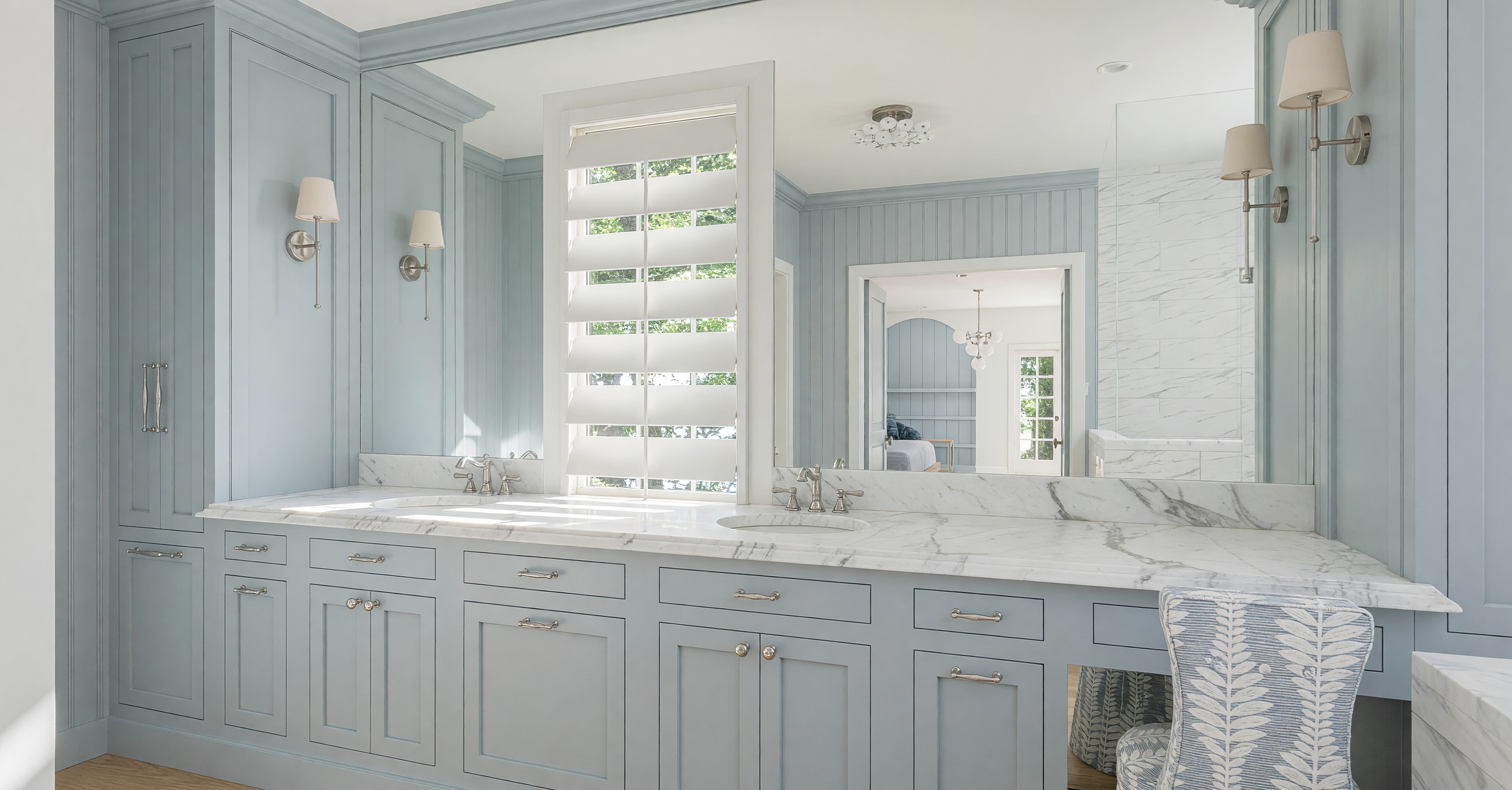 A bathroom vanity with a marble countertop, two sinks, silver faucets, light blue cabinetry, and modern wall sconces on either side of a large mirror. A window with white louvers allows natural light to enter, and a reflection in the mirror shows a glimpse of an adjoining room.