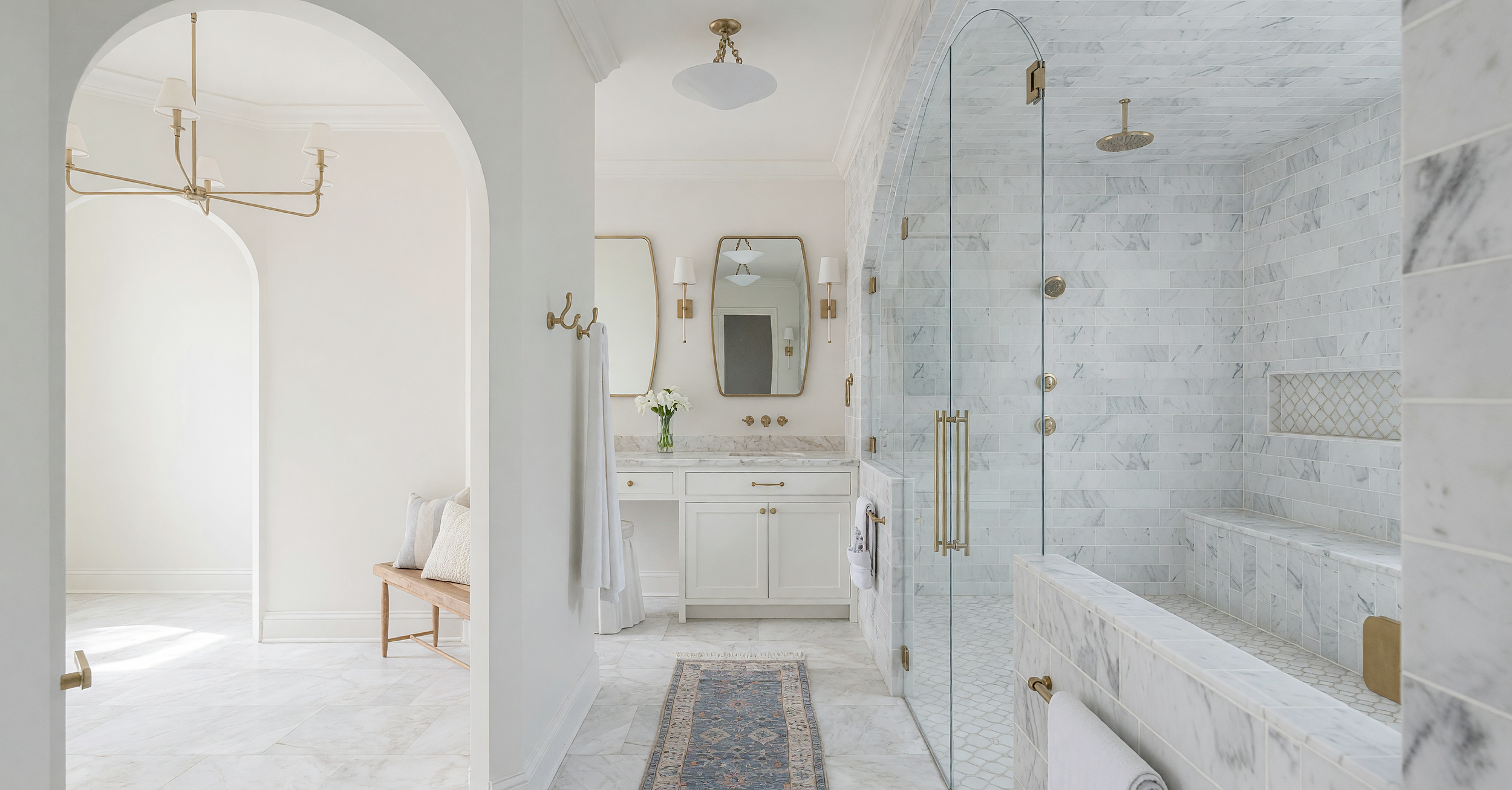 A luxurious bathroom with white marble tiles, a glass-enclosed shower with brass fixtures, a white vanity with gold handles, and minimal decor including mirrors, lamps, and a vase of flowers.