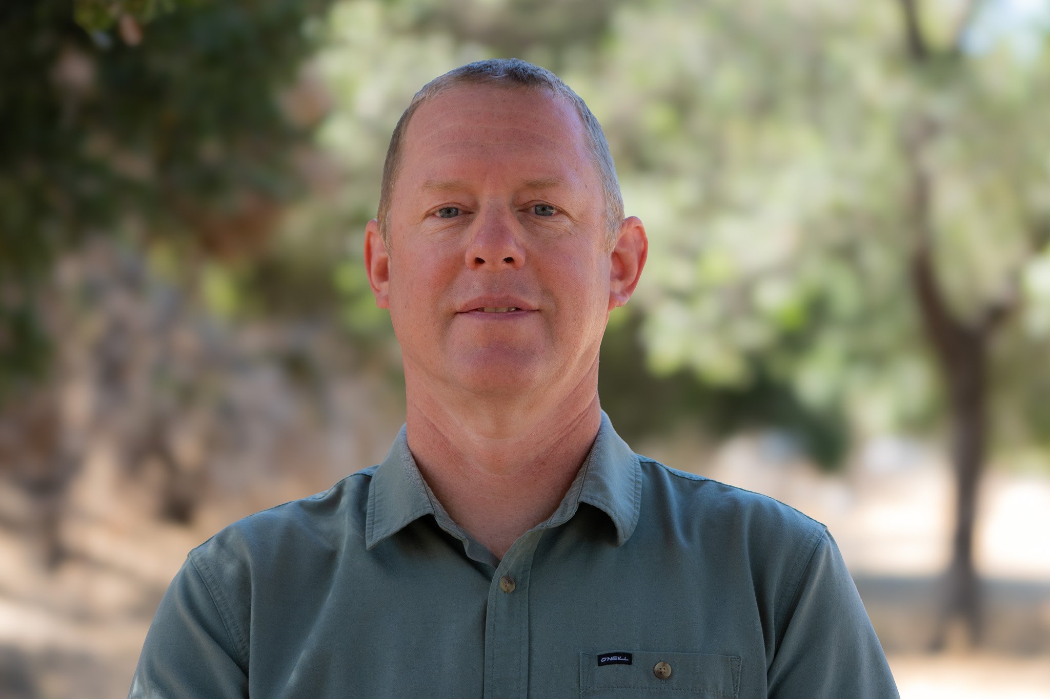 A middle-aged man with short hair and blue eyes, wearing a gray button-up shirt, standing outdoors with blurred green trees in the background.