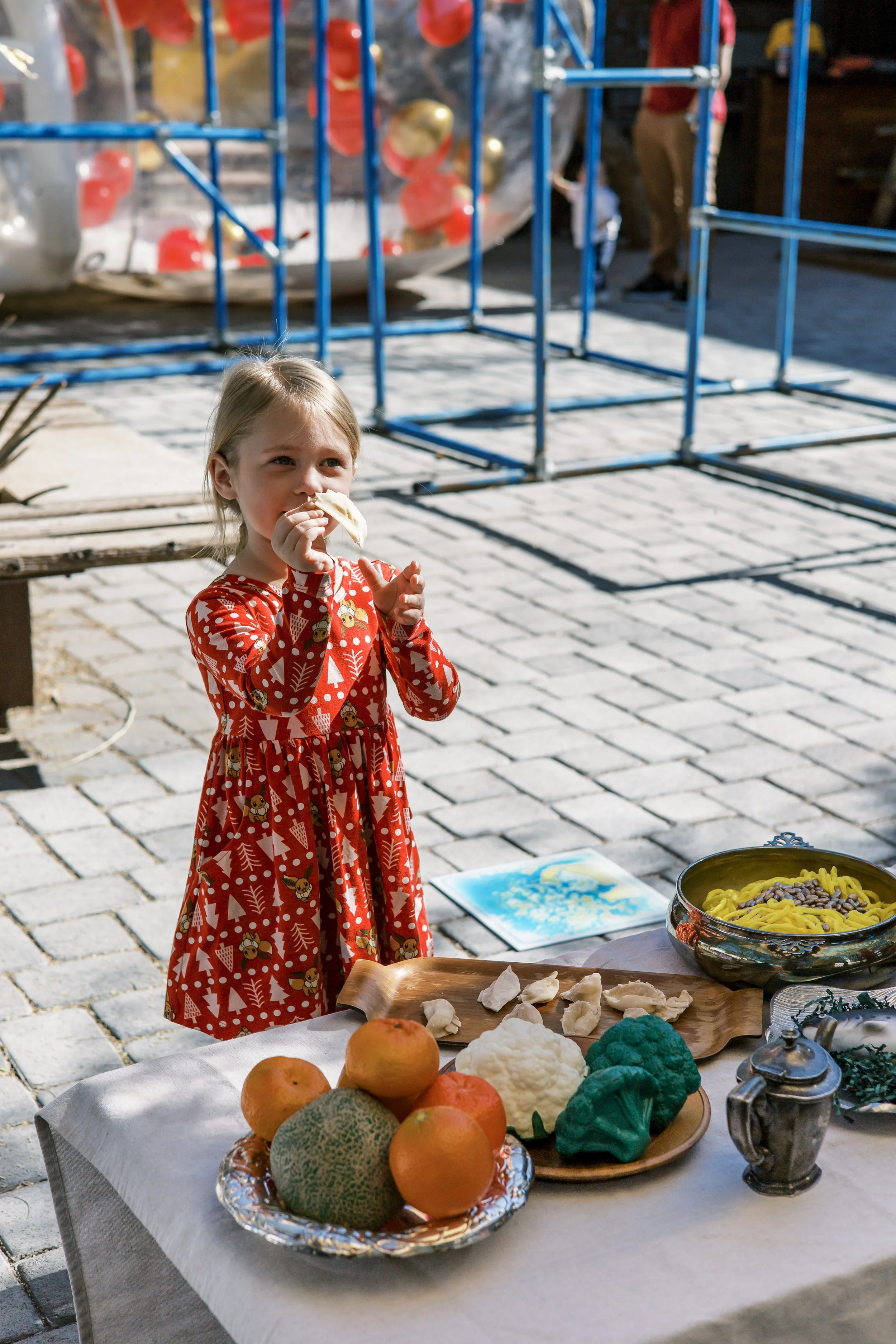 A young girl in a red festive dress with reindeer and Christmas trees, eating food at an outdoor table with fruit and dishes, with a playground or climbing structure in the background.