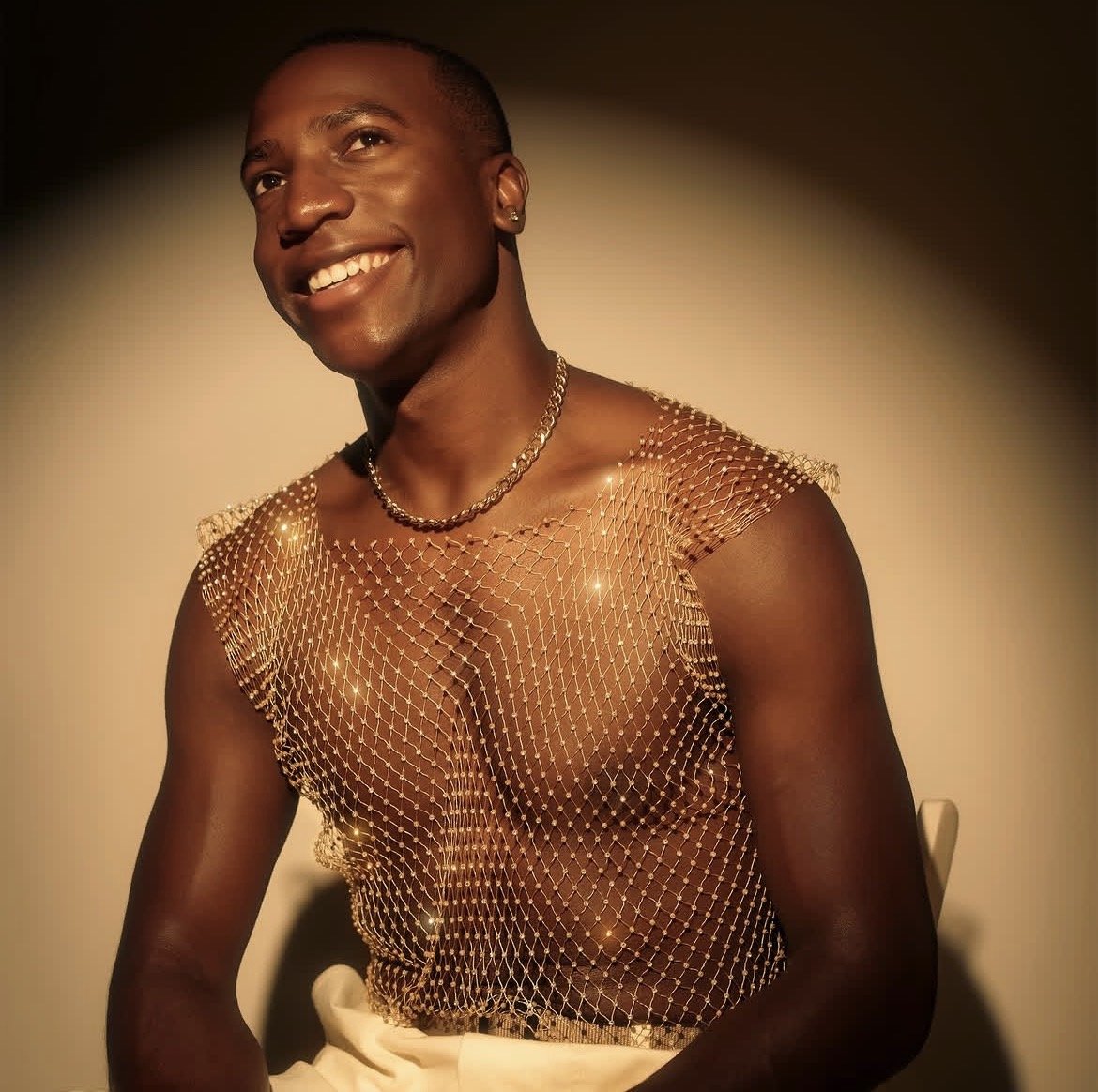 A smiling person with short hair wearing a sheer, embellished top and a silver chain necklace, posing against a neutral background.
