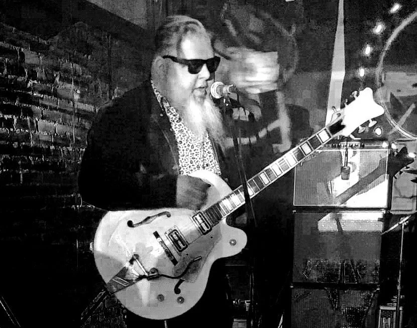 A man with sunglasses and a beard playing a semi-hollow electric guitar and singing into a microphone in a music venue.