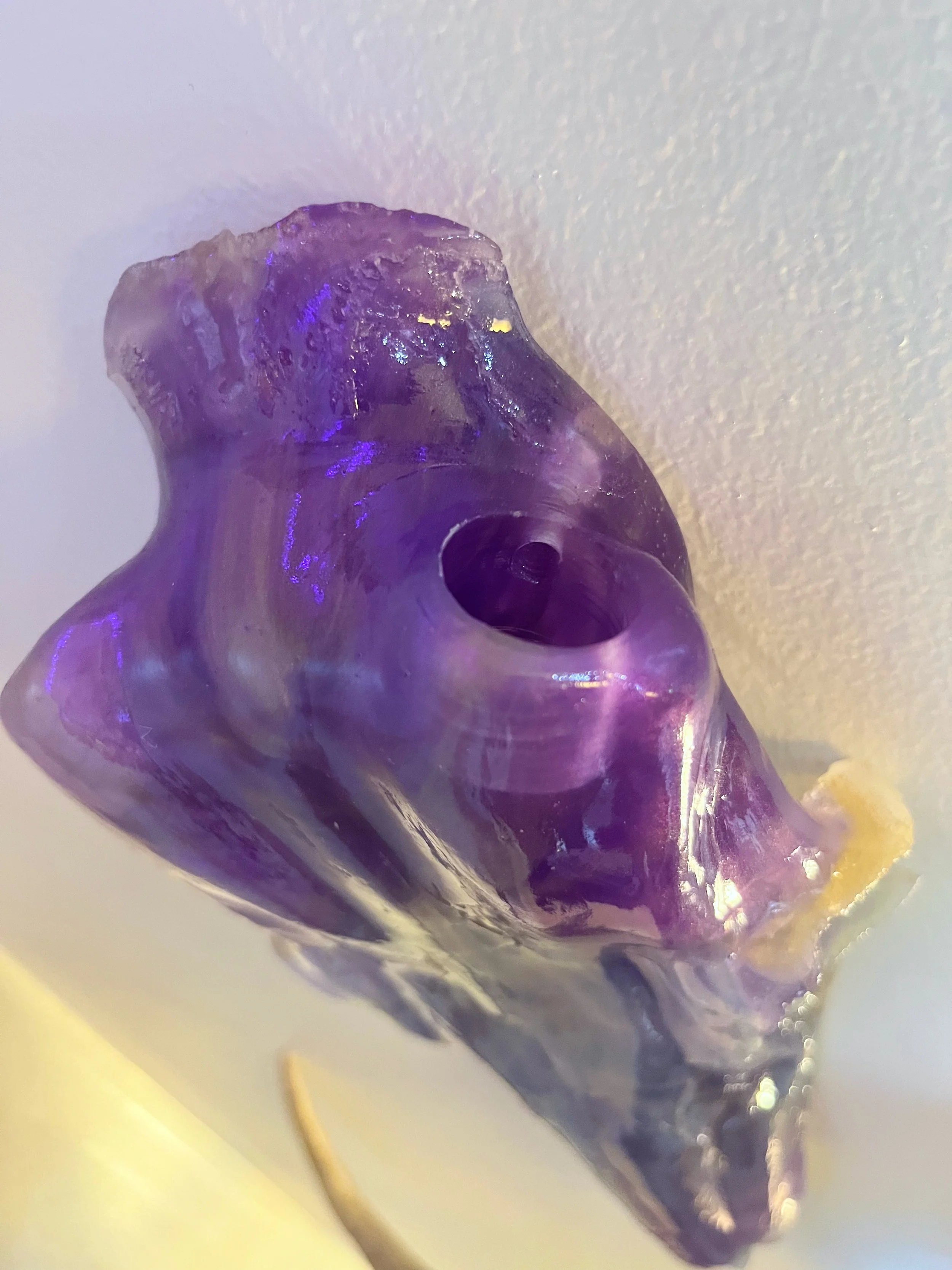 Close-up image of a purple, translucent, glass skull with a hole at the top, placed on a white surface.