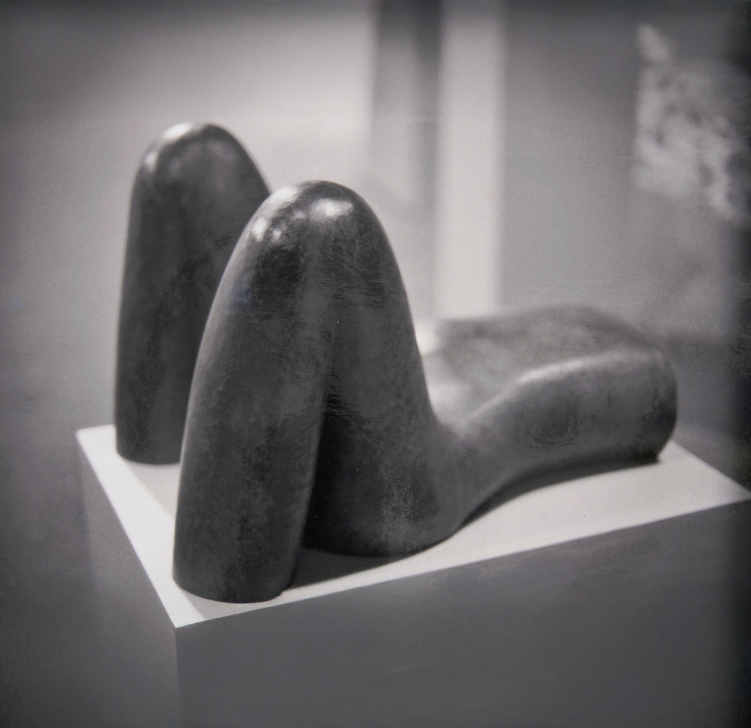 A black sculpture of a reclining figure with bent knees and arms, displayed on a white pedestal in a gallery setting.